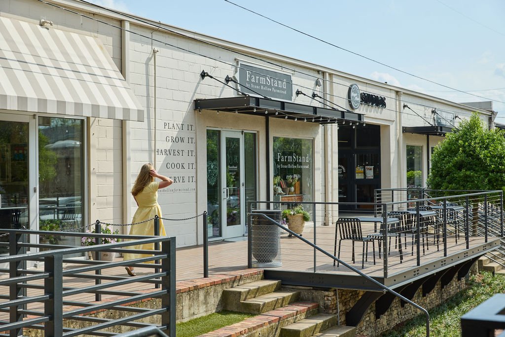 A woman in a yellow dress walking on an outdoor patio of a store called FarmStand by Stone Hollow Farmstead, with black chairs and tables, potted plants, and a large storefront window with a striped awning.