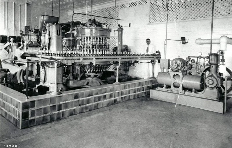 Black and white photo of an industrial machine in a factory with workers, pipes, and machinery setup on a tiled platform.
