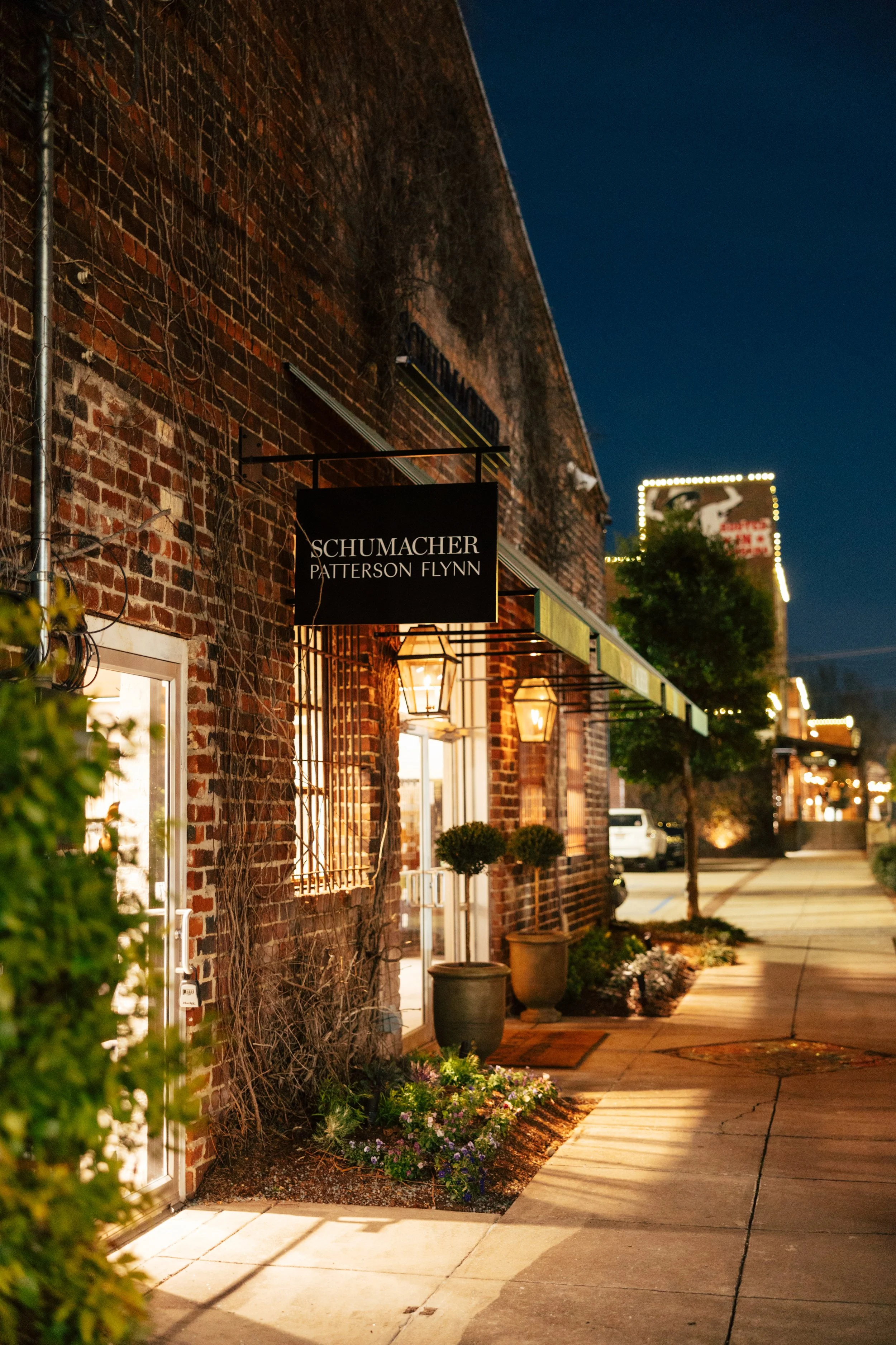 Nighttime street view of a brick building with illuminated porch lights, potted plants, and a sign that reads 'Schumacher Patterson Flynn'.