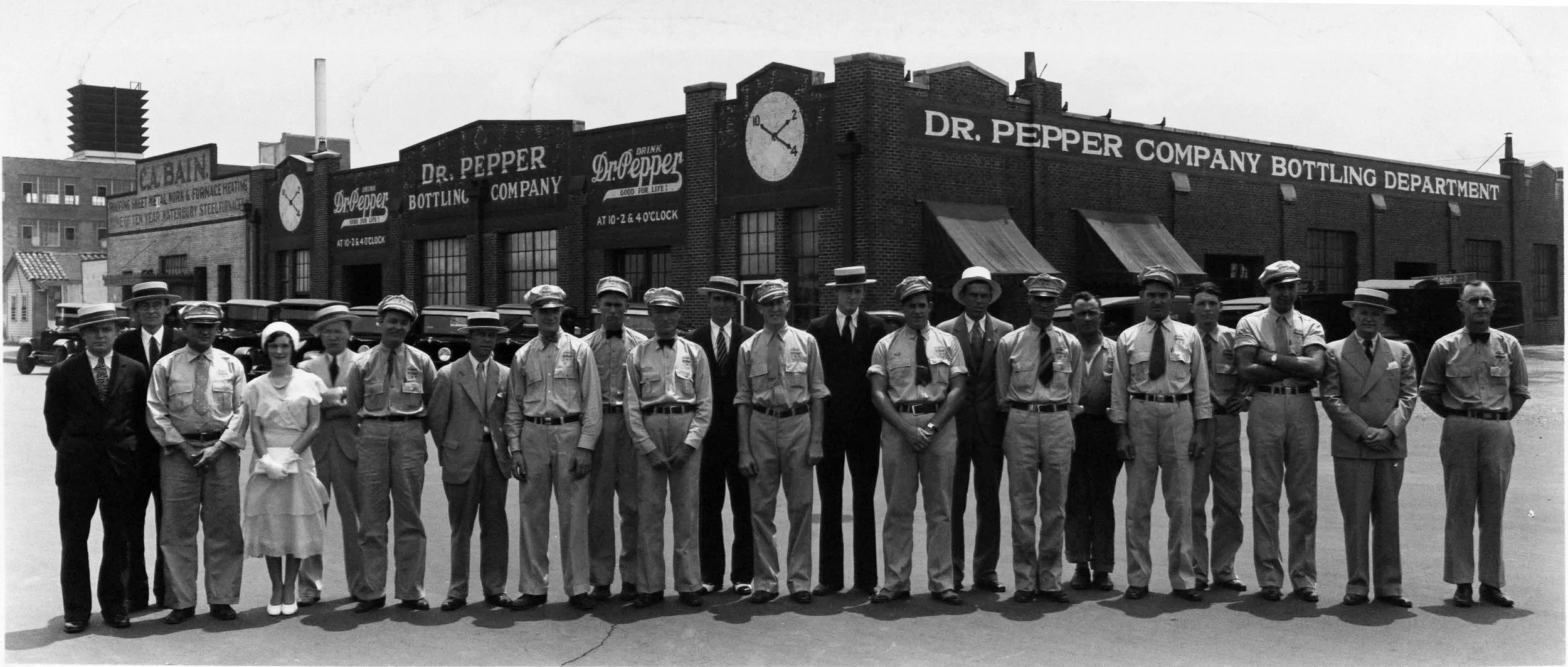 Black and white photograph of a group of men and women standing in front of a building labeled Dr. Pepper Bottling Company. The building has signs indicating bottle filling times and a large clock. The people appear to be workers and managers, dressed in period clothing, including some in uniform and others in formal attire with hats.