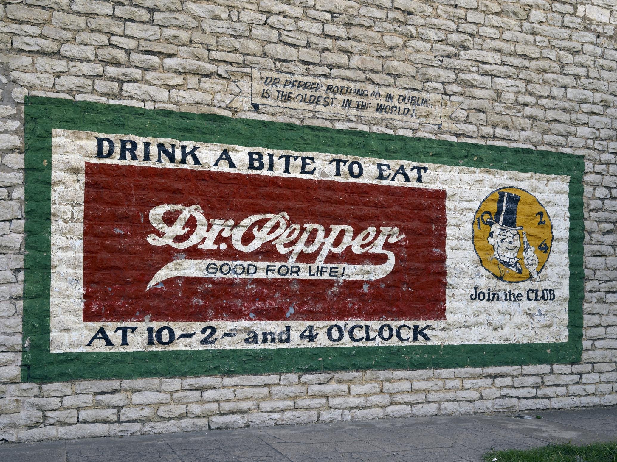 A painted advertisement on a brick wall for Dr. Pepper. It features the slogan 'Drink a Bite to Eat' and mentions the drink is good for life. The ad indicates the establishment is open from 10 AM to 2 AM and 4 PM. There is a circular logo with a cartoon man wearing a tall hat and monocle, with the text 'Join the Club' beside it.