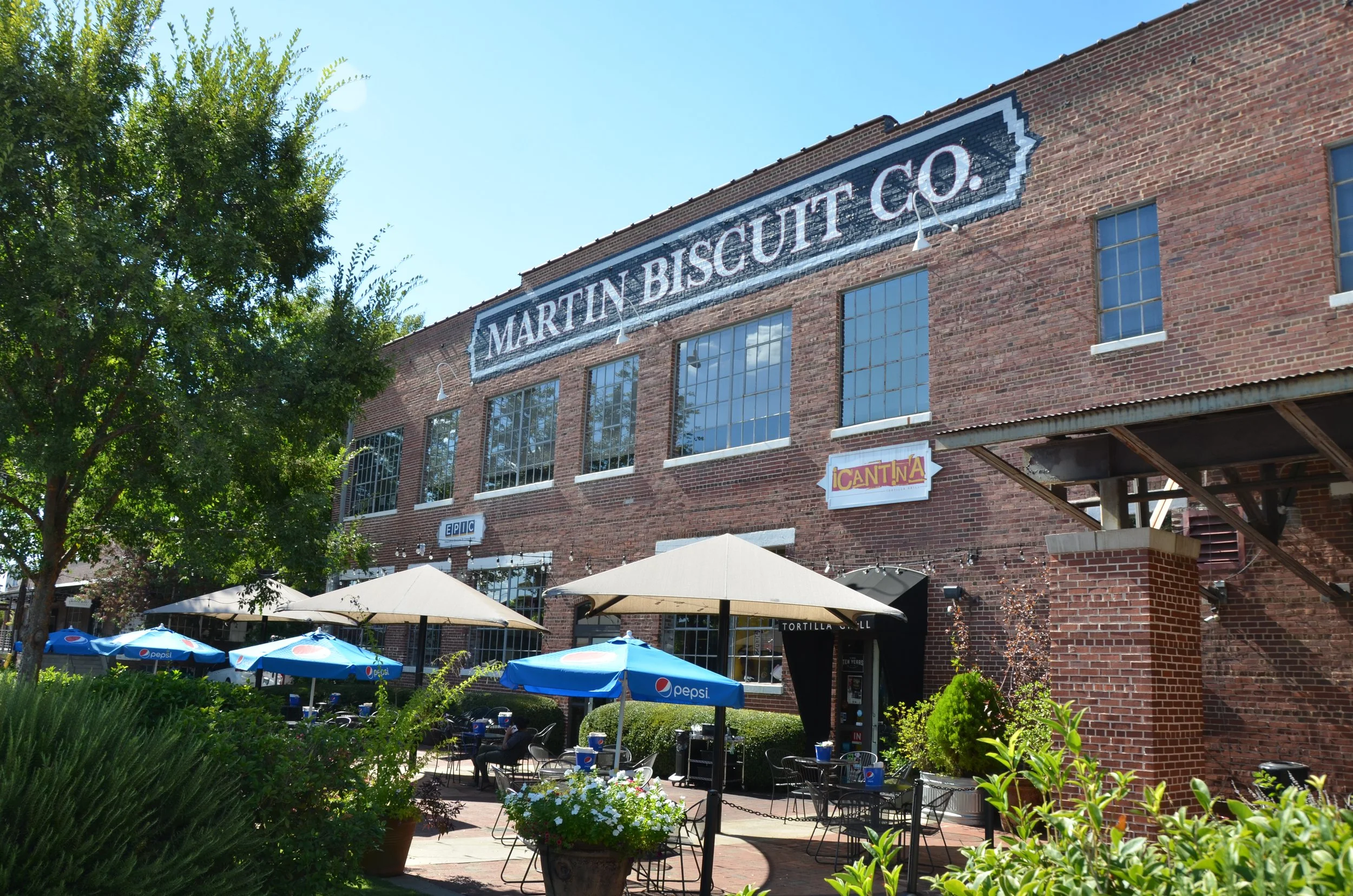 Outdoor patio area with umbrellas and tables in front of a brick building with large windows. The building has signage that reads 'Martin Biscuit Co.' and smaller signs for 'Epic' and 'Cantina.' There is a large green tree on the left side and various plants in the foreground.