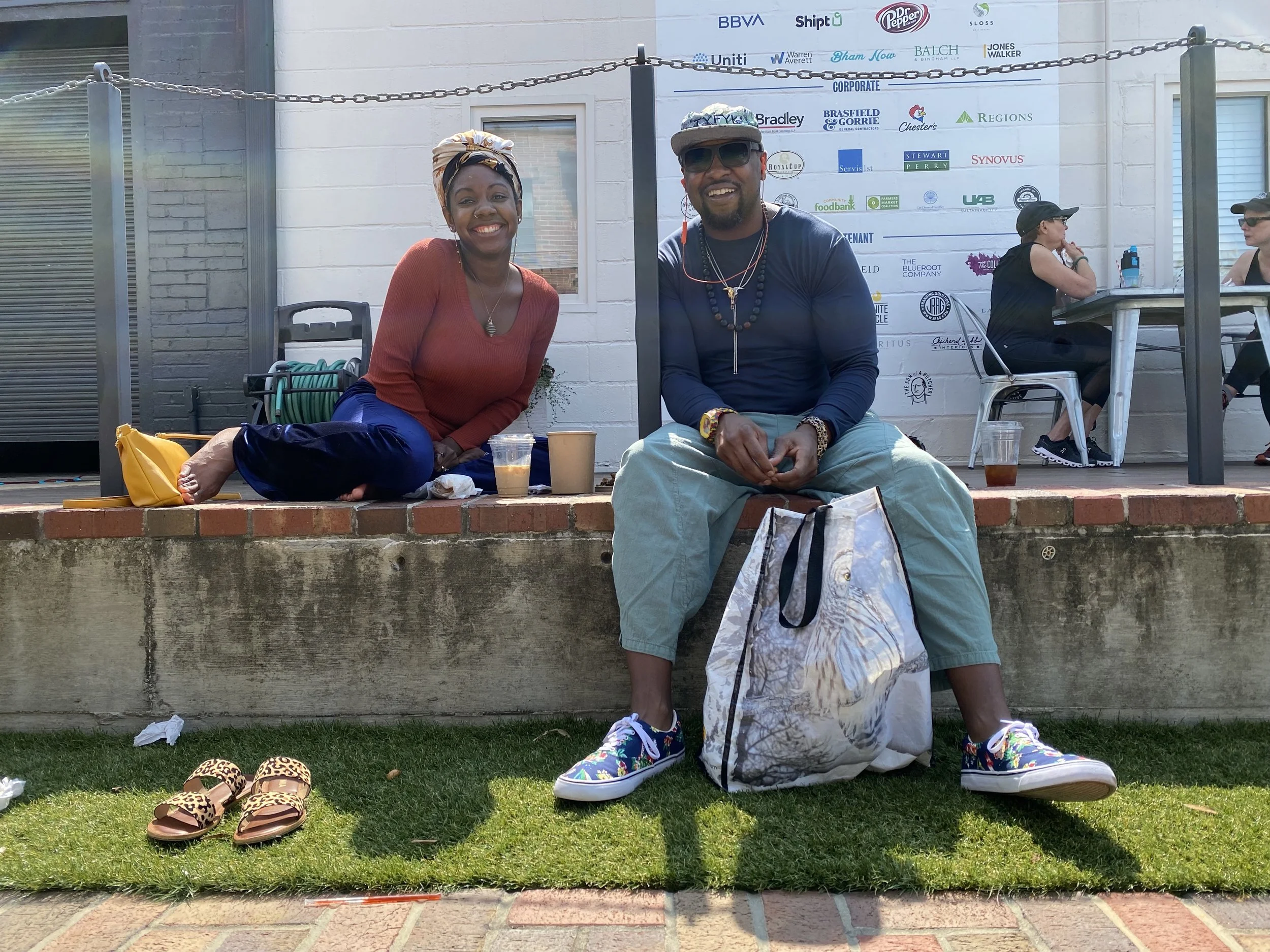 Two people sitting on a brick ledge outdoors, smiling at the camera, with a white building background and a large promotional banner behind them. One woman is wearing a headscarf, red long-sleeve shirt, and blue pants, with sandals on the grass nearby. The man wears sunglasses, a baseball cap, a long-sleeve navy shirt, and light green pants, with a large bag resting on his lap.