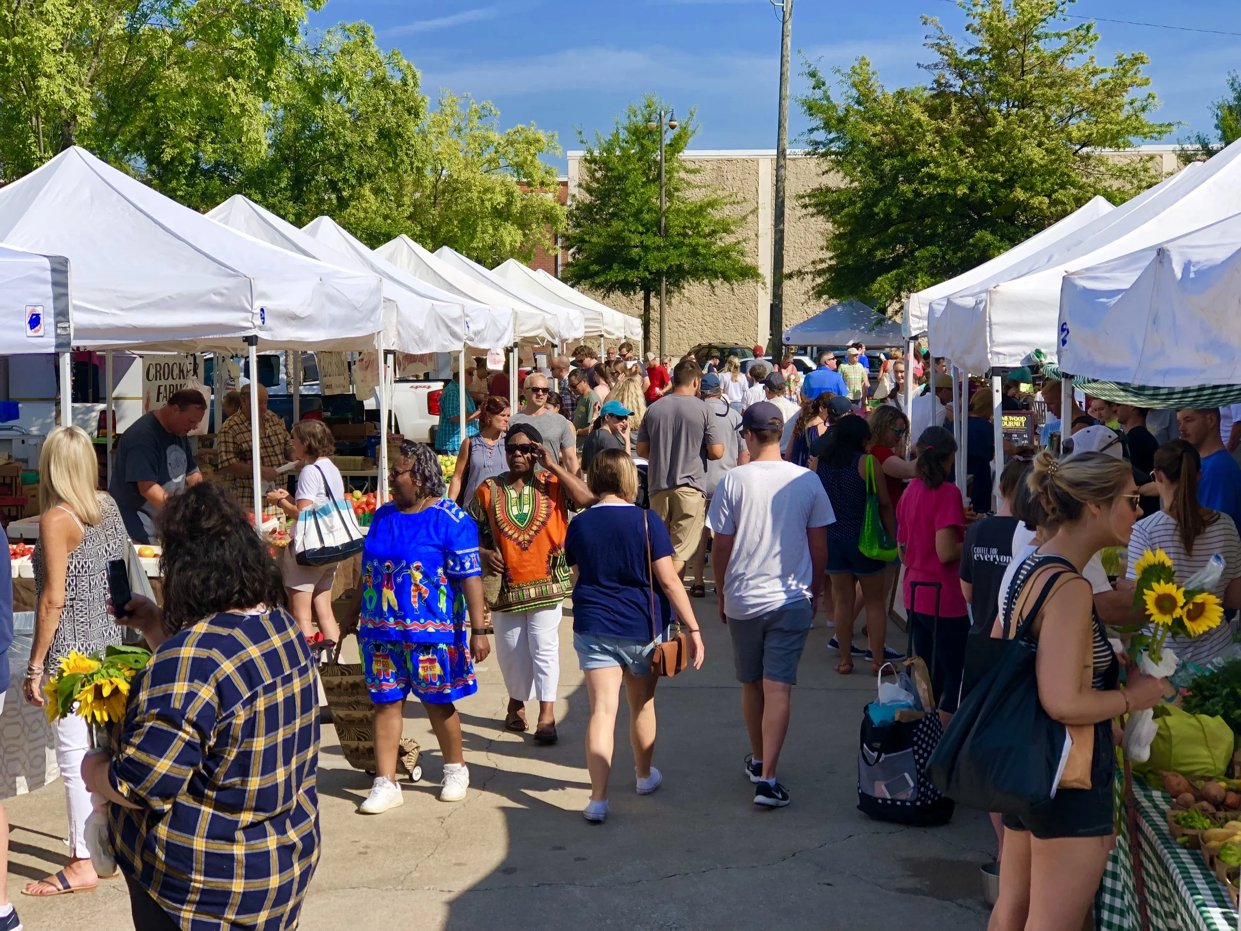 People shopping at an outdoor market with white tents, trees, and a building in the background on a sunny day.