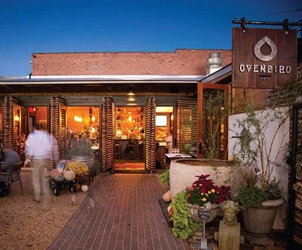 Exterior view of Ovenbird restaurant at dusk with patio seating, potted plants, and a sign with the restaurant's name.