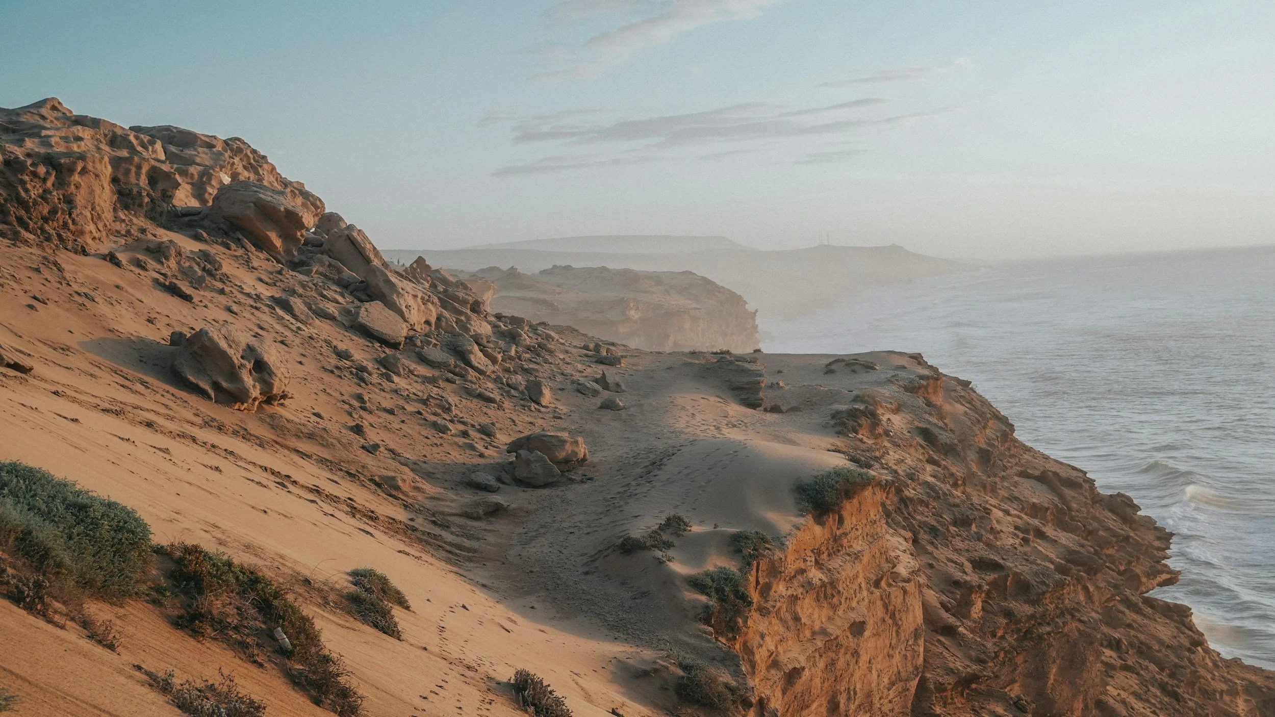 Sandstone cliffs and rugged terrain along a coastline with ocean waves, under a cloudy sky during sunset.