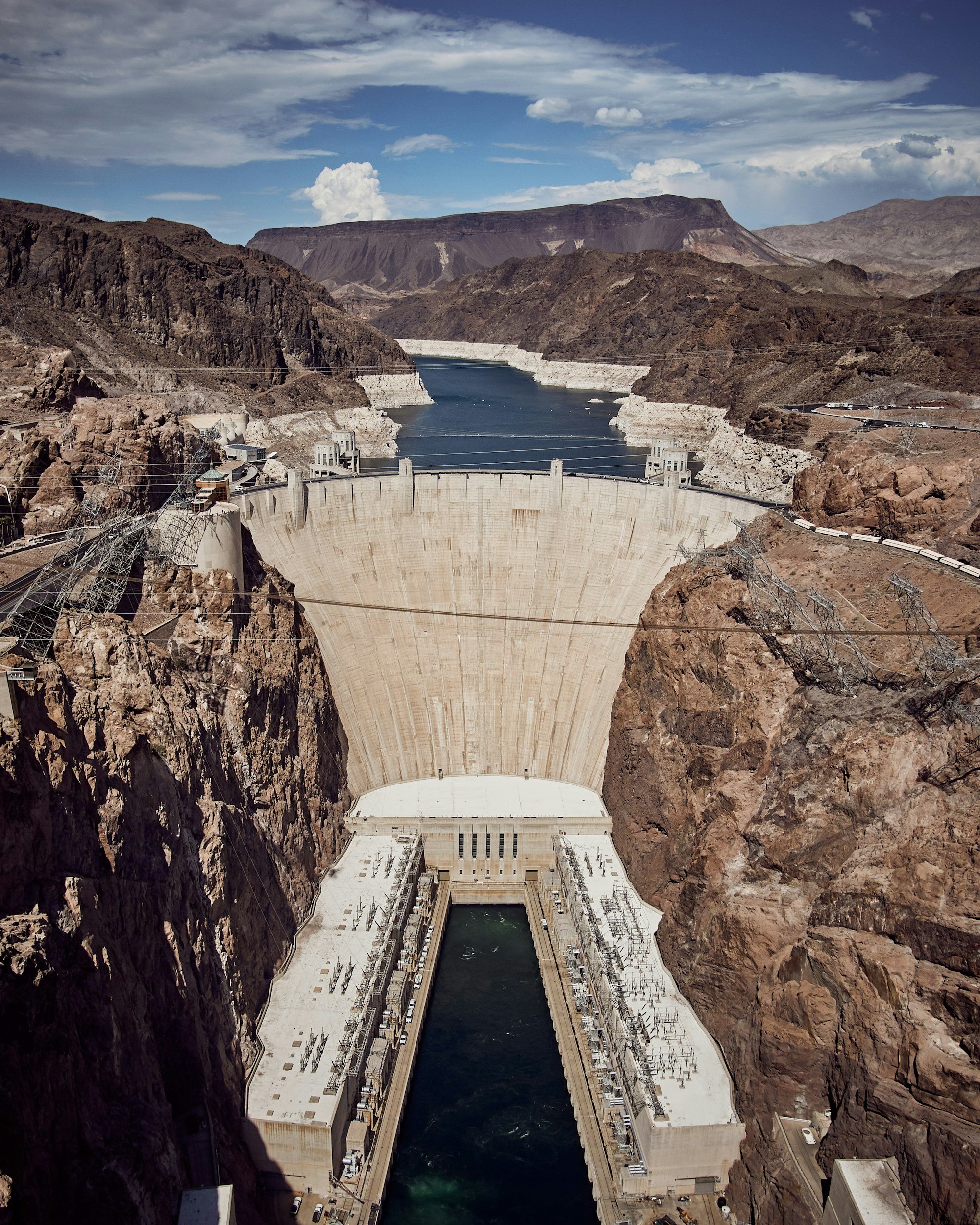 The image shows the Hoover Dam, an iconic concrete arch-gravity dam located on the border between Nevada and Arizona, in a mountainous desert landscape under a partly cloudy sky.