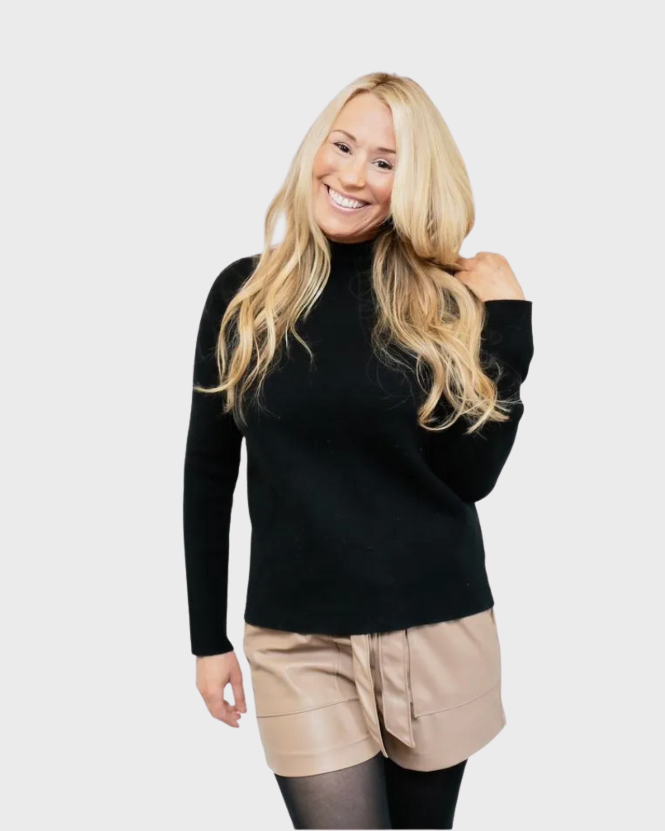 A woman with long blonde hair wearing a black long-sleeve top, beige shorts, and black tights, smiling and touching her hair.