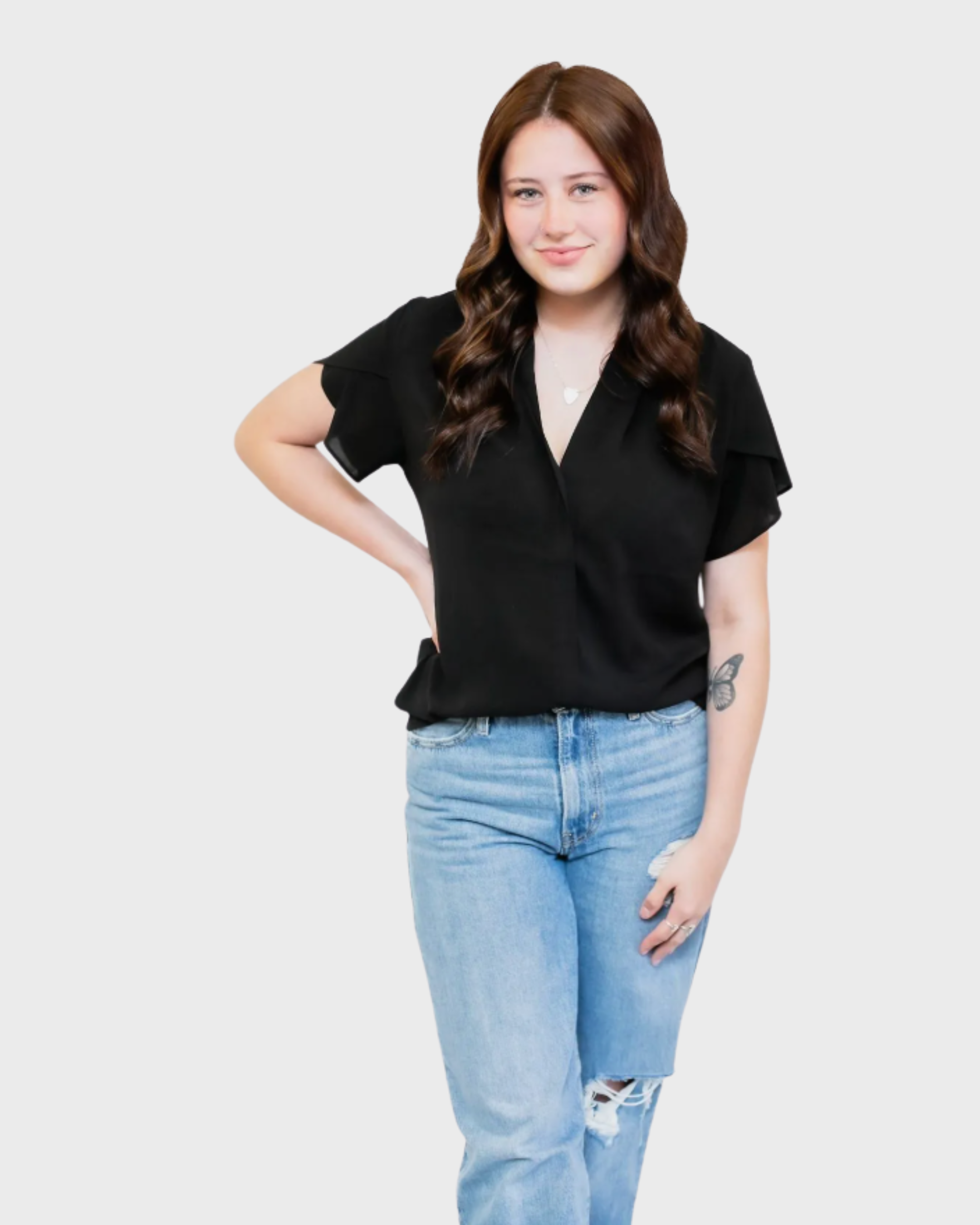 Young woman with brown wavy hair wearing a black blouse, light blue ripped jeans, standing against a plain background.