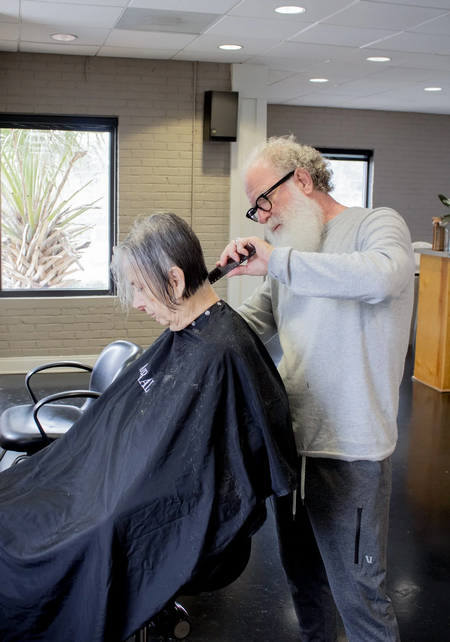 An older man cutting a woman's hair in a salon, with hair clippings on her cape and hairdresser focused on his work.