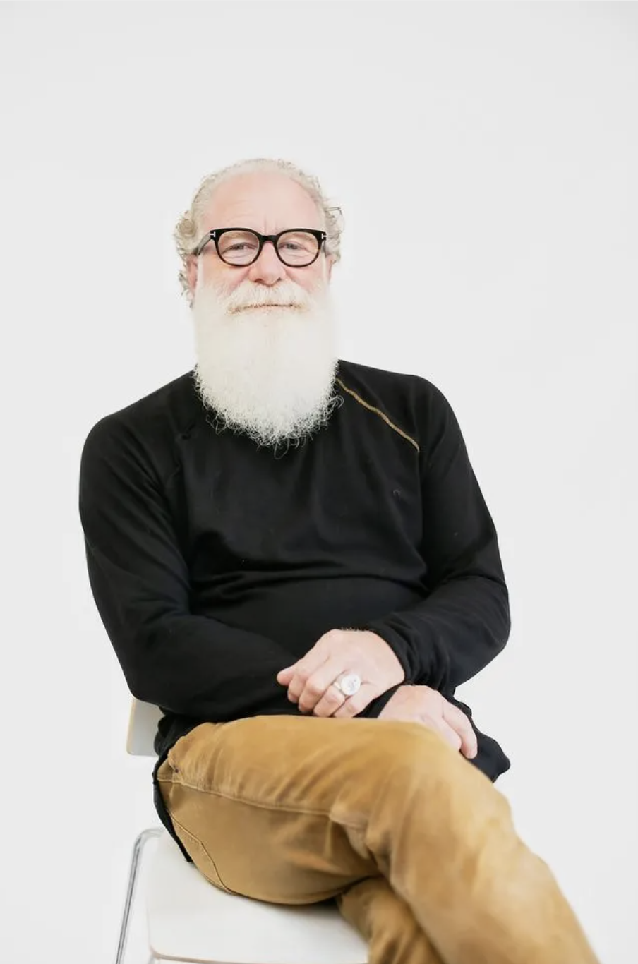 An older man with a white beard and glasses, wearing a black sweater and tan pants, sitting on a white chair against a plain white background.