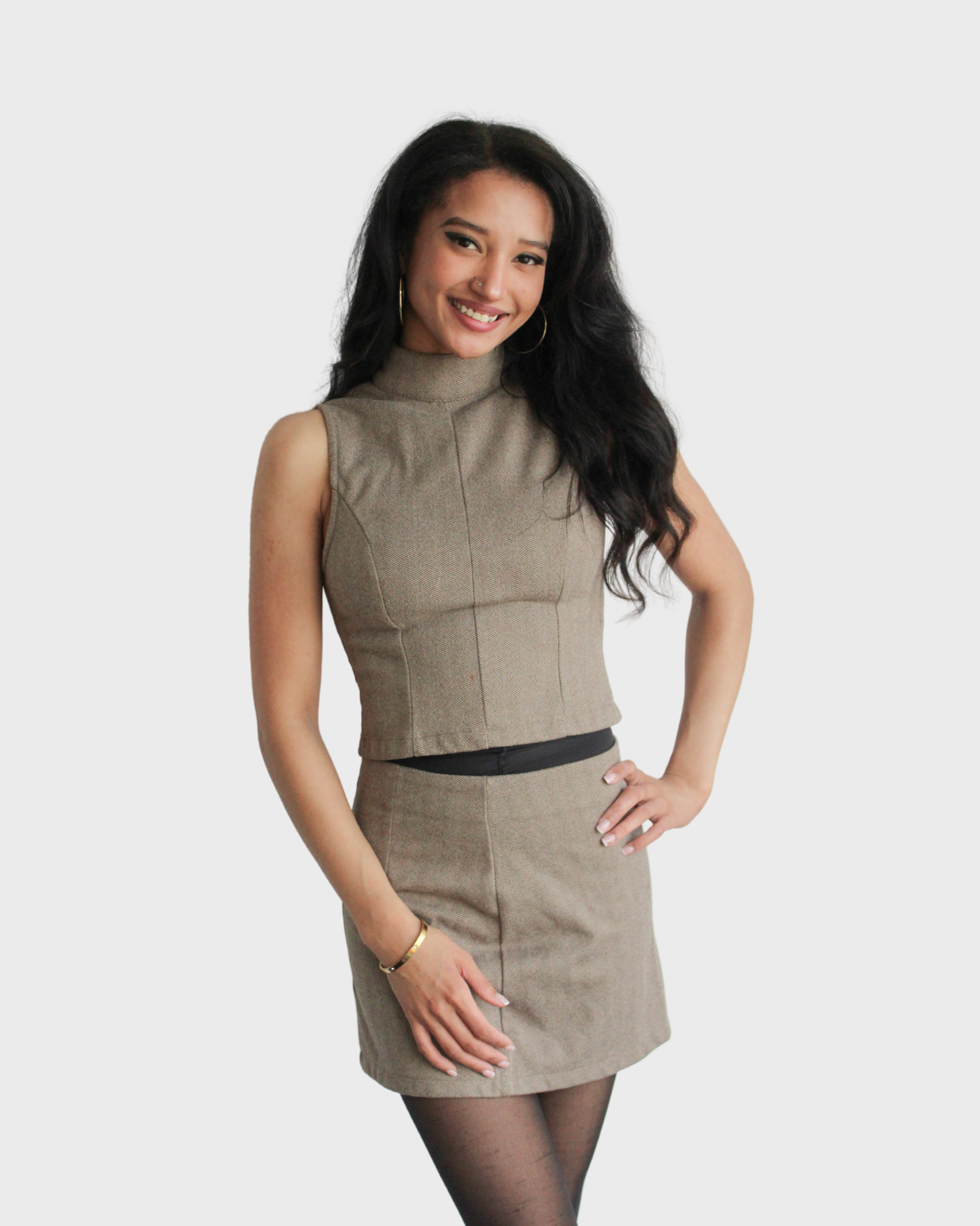 Smiling woman with dark wavy hair, wearing a beige sleeveless turtleneck top and matching skirt, black tights, standing with one hand on her hip against a plain light gray background.