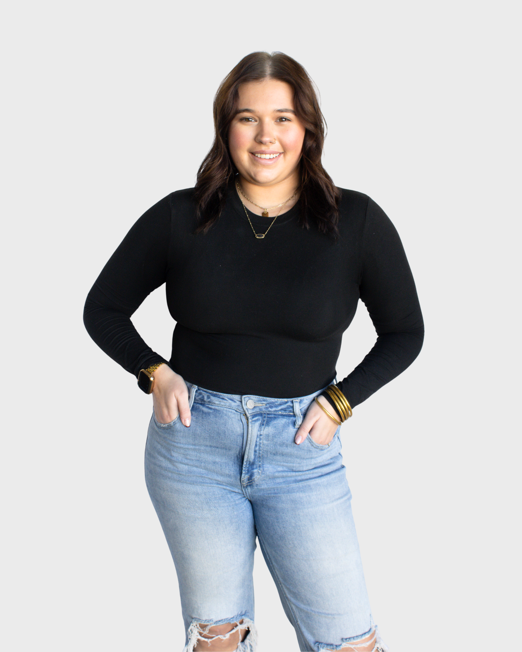 Young woman with shoulder-length brown hair, wearing a black long-sleeve top, light blue ripped jeans, gold jewelry, and a smartwatch, smiling and standing against a plain white background.