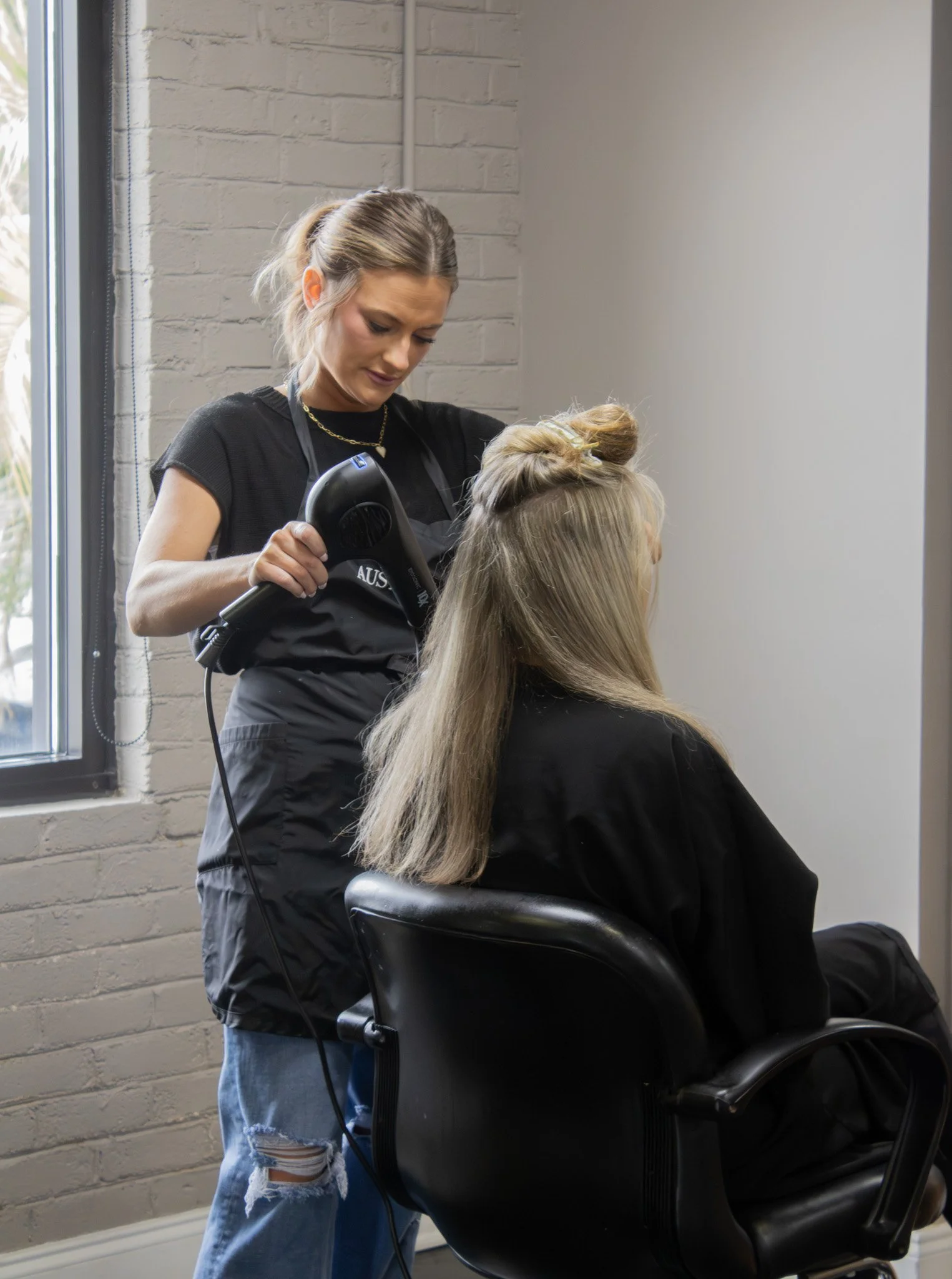 A hairstylist blow-drying a woman's hair in a salon, with the woman seated in a black chair near a window.