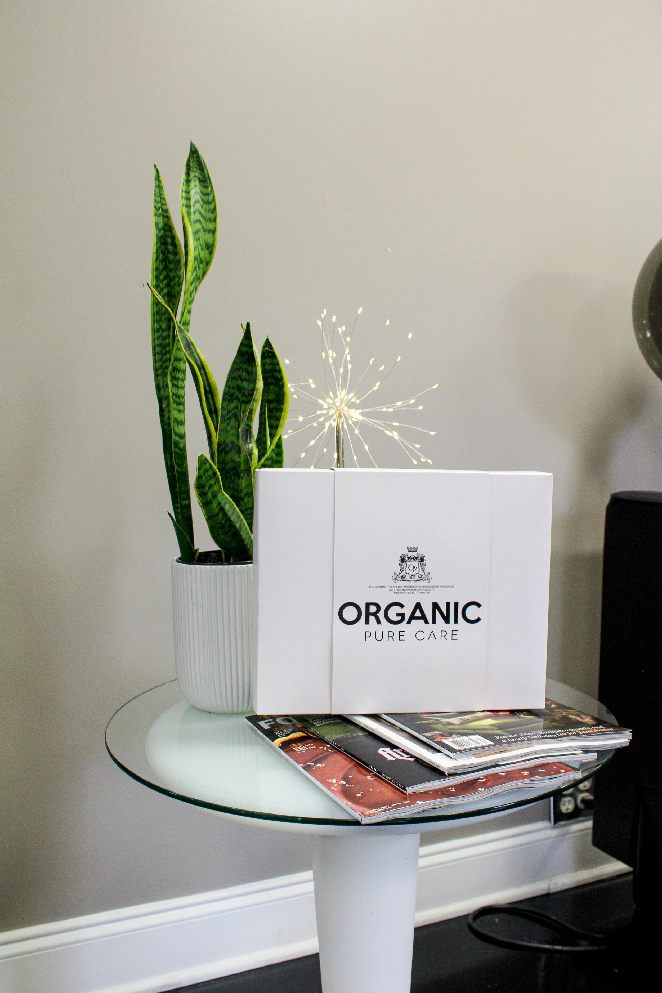 A white round table holding a white potted snake plant, a white box labeled 'Organic Pure Care,' and several magazines.