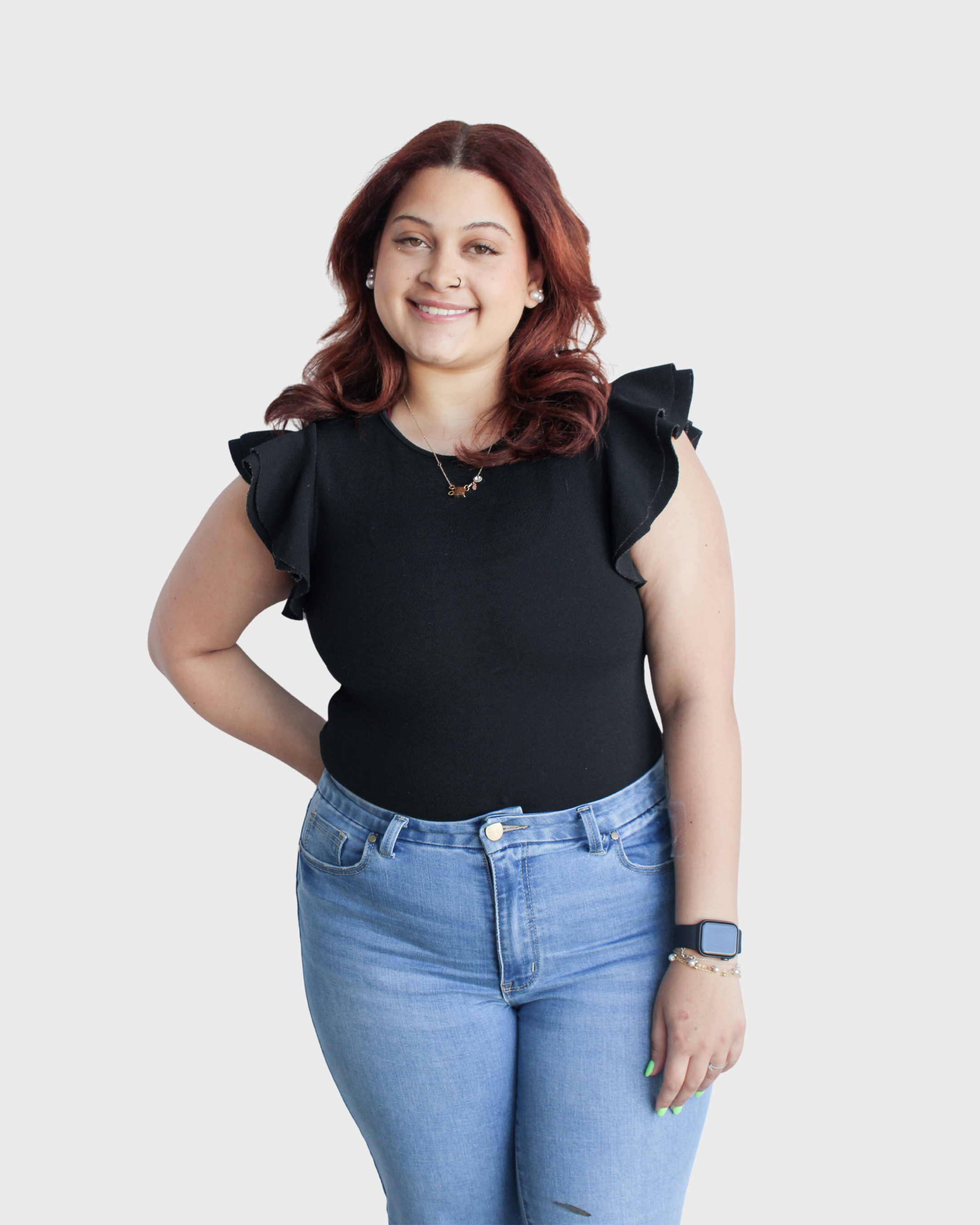 A woman with shoulder-length reddish-brown hair wearing a black top with ruffled sleeves, light blue jeans, an Apple Watch, and jewelry, standing against a light background and smiling.
