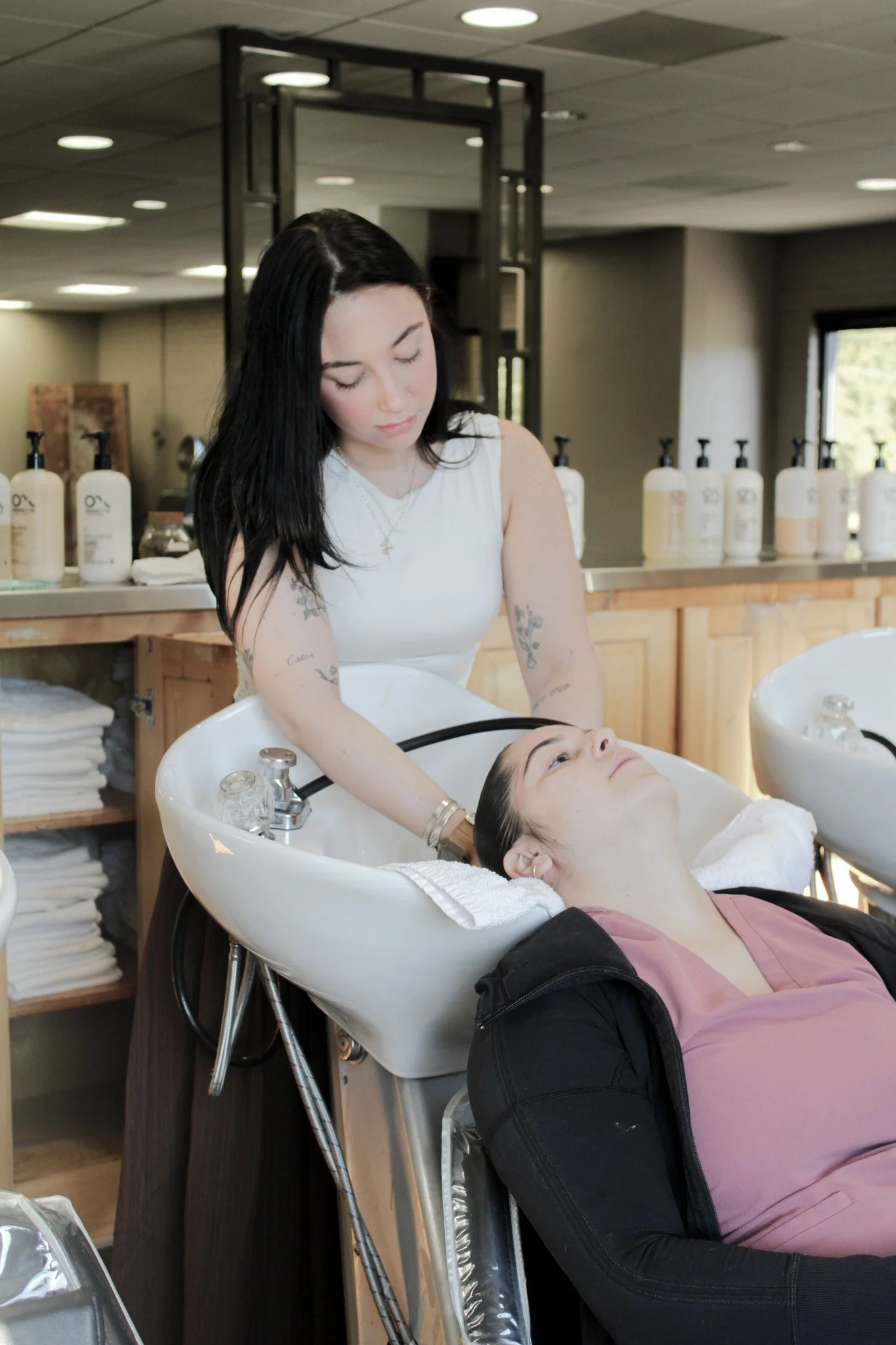 A woman receiving a hair wash at a salon with black hair and tattoos, lying back in a white salon sink with another woman washing her hair.