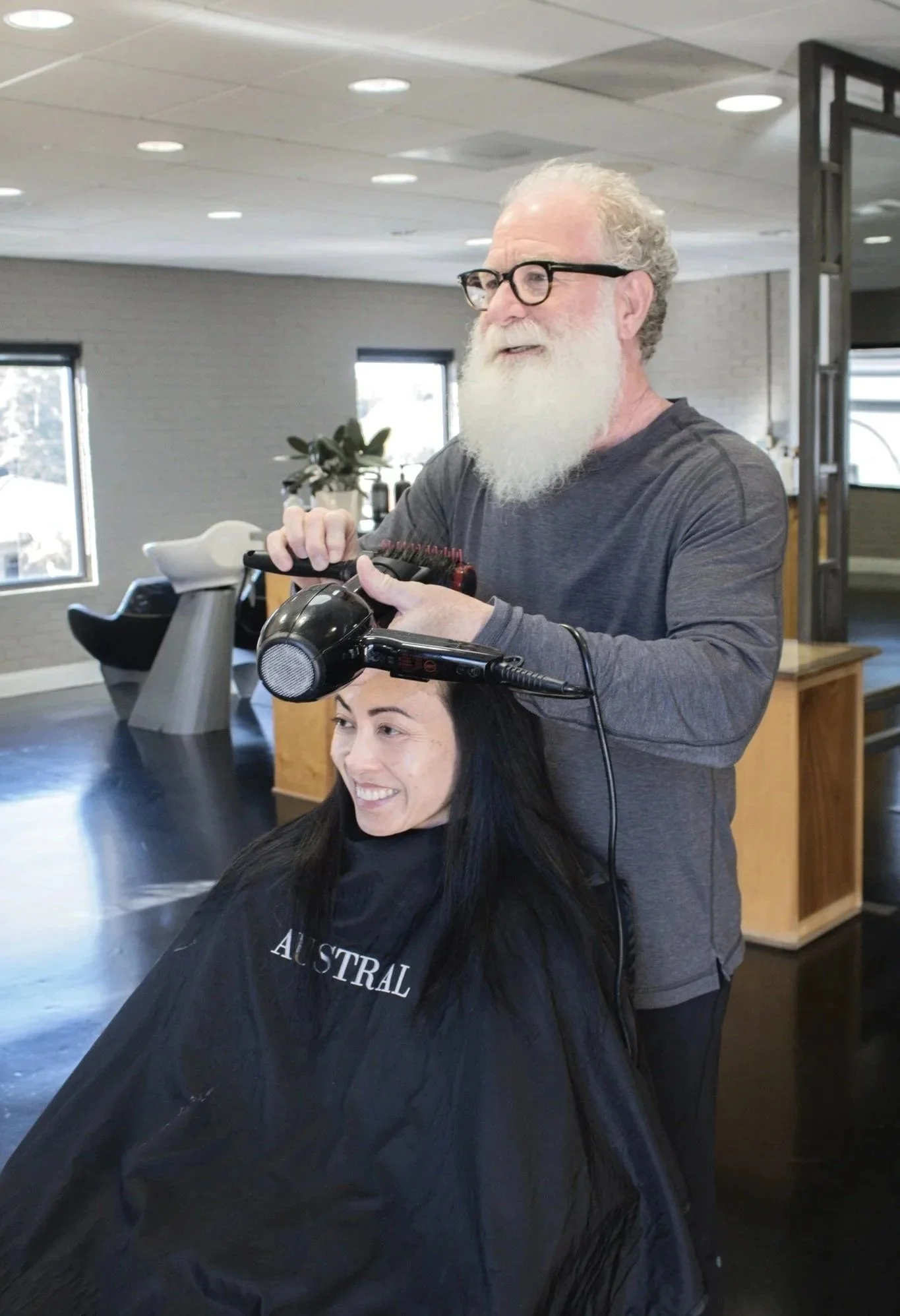 A man with glasses and a gray beard is using a hot air brush to style the hair of a woman with dark hair, who is seated and smiling in a hair salon.