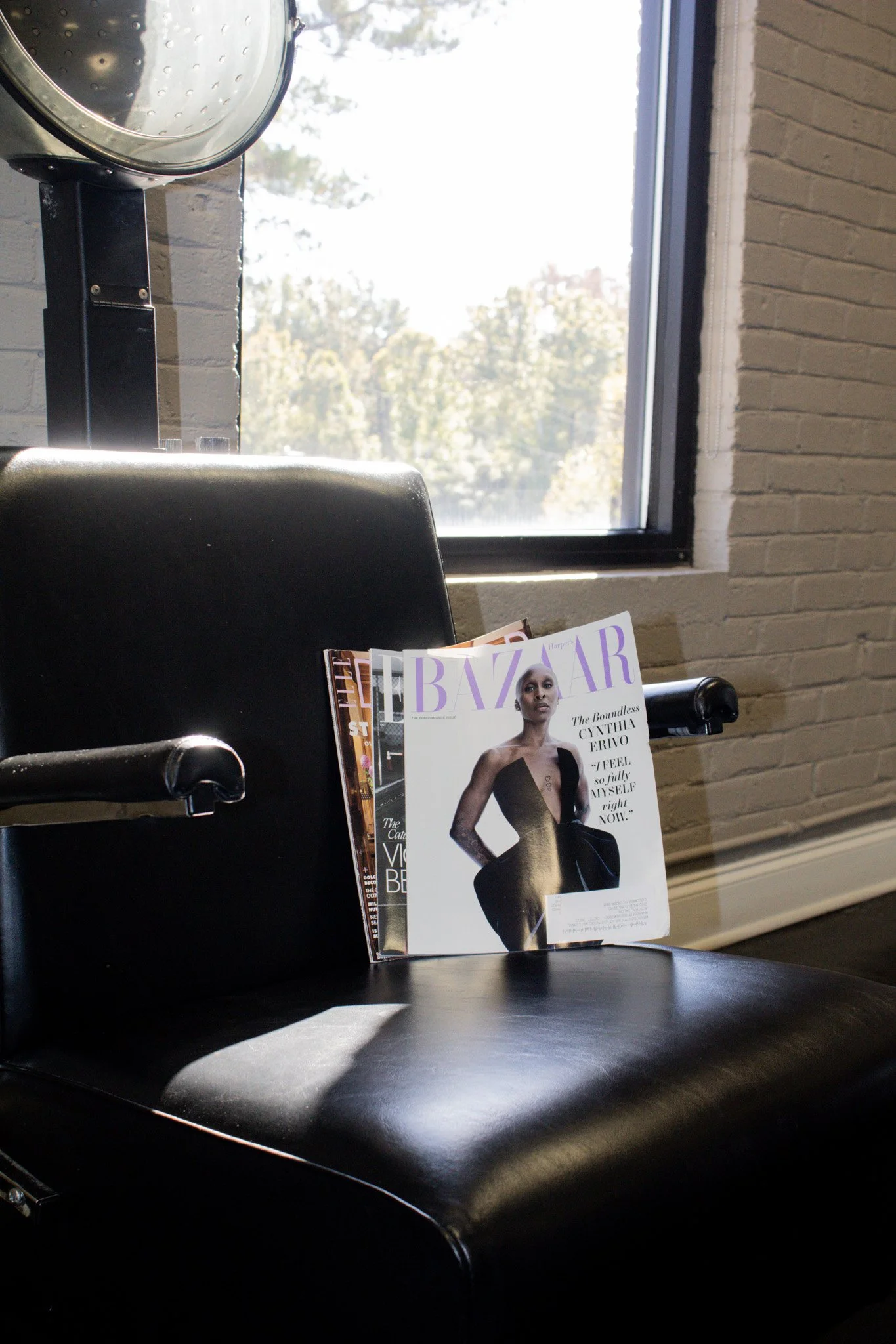 A black salon chair with an open Harper's Bazaar magazine on the seat near a window with a white brick wall background.