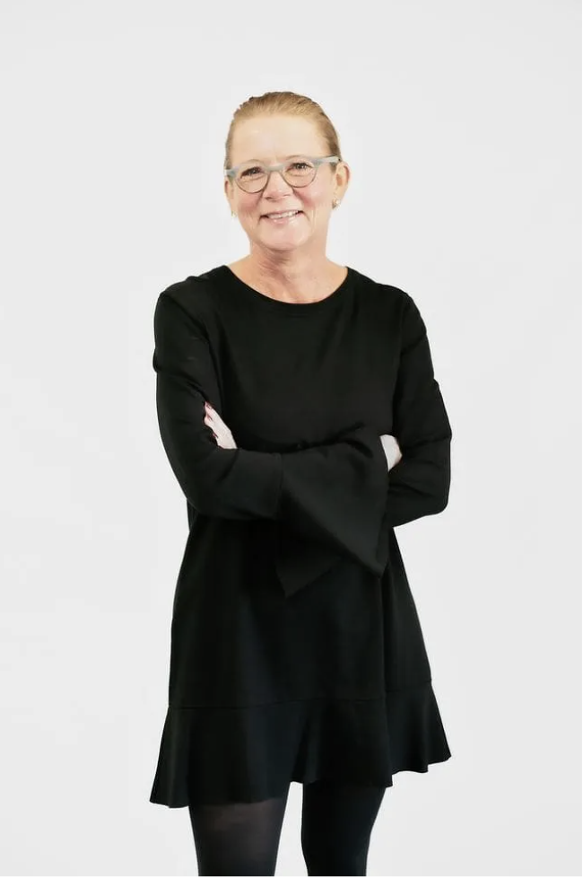 An older woman with light skin, glasses, and red hair tied back, smiling with arms crossed, wearing a black dress and black tights against a plain white background.