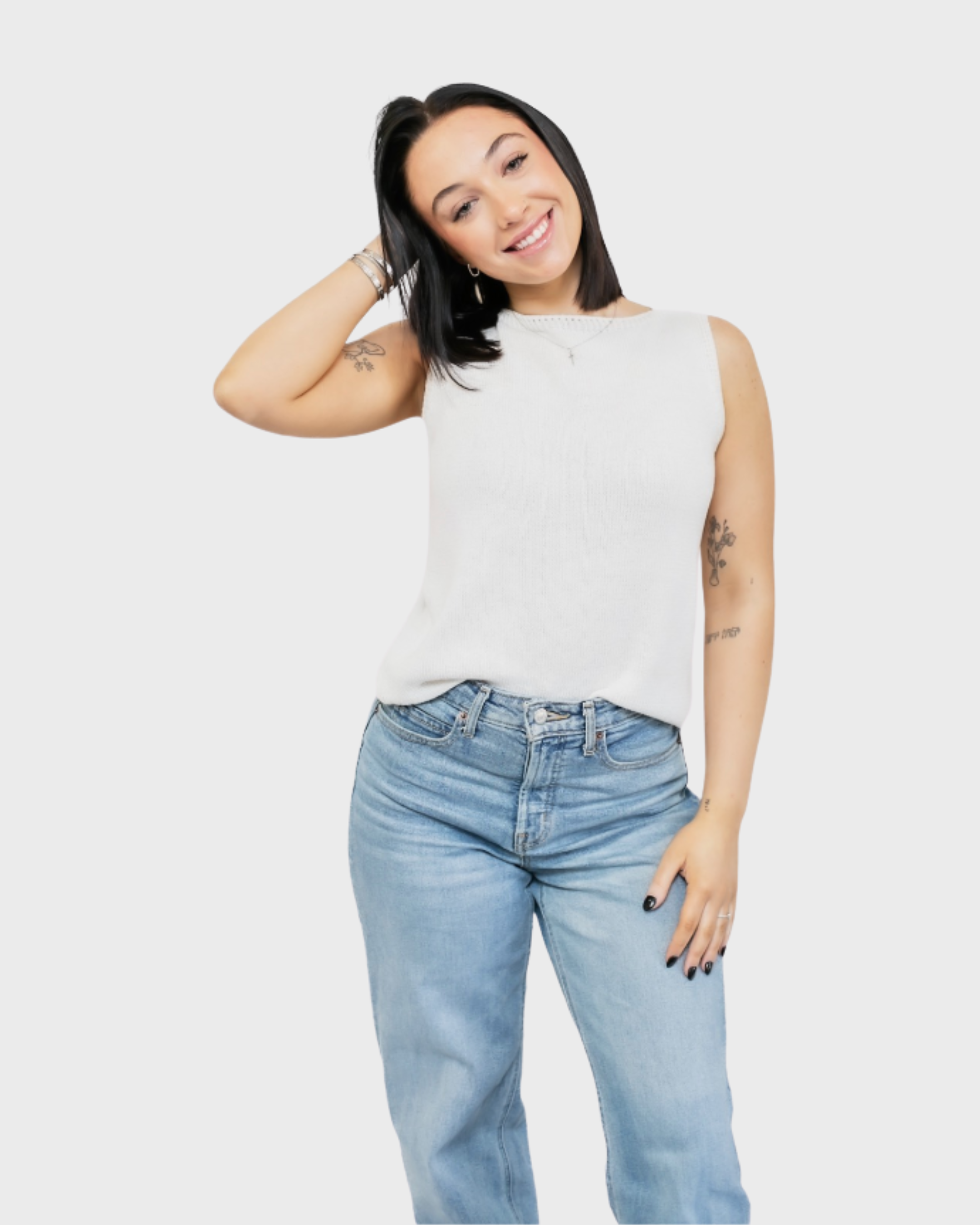 A woman with black hair and tattoos on her arms, smiling and posing with her hand on her head, wearing a white sleeveless top and light blue jeans.