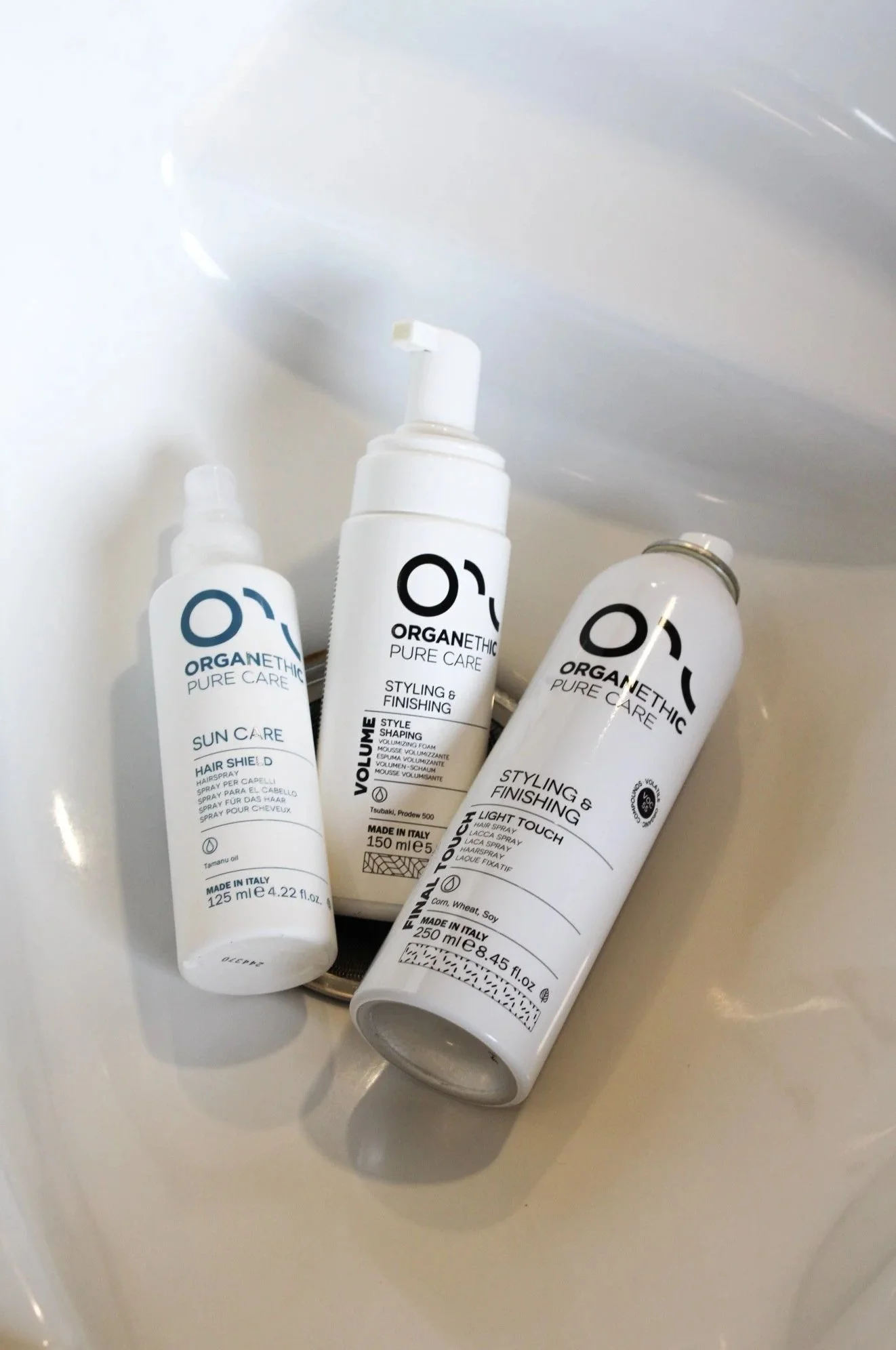 Three bottles of Organic Pure Care styling and finishing products resting on a sink or basin, including a volume foam, light touch spray, and hair shield spray.