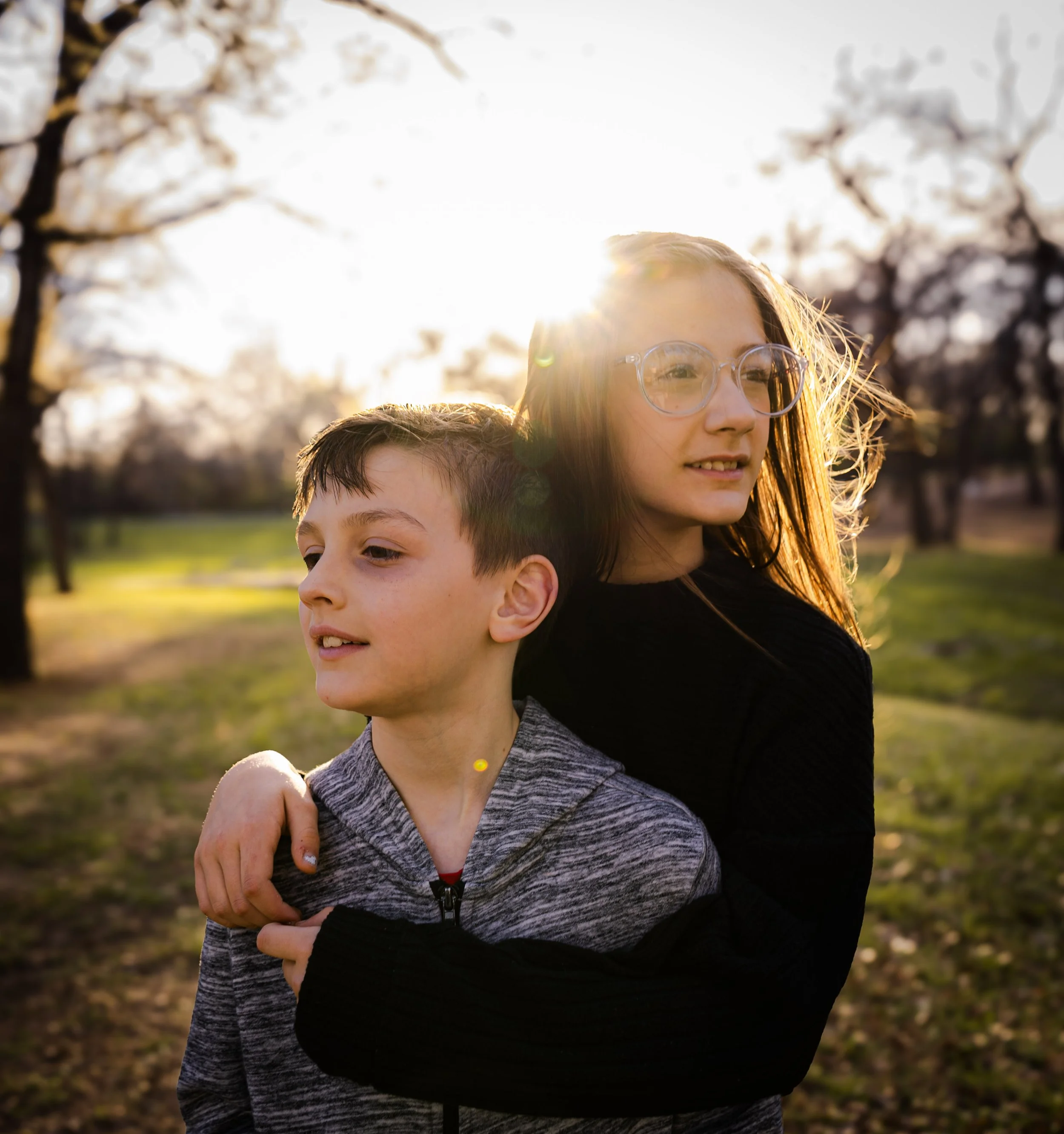 A young girl with glasses and long hair, and a young boy with short hair, standing outdoors in a park during sunset, with trees and grass in the background.