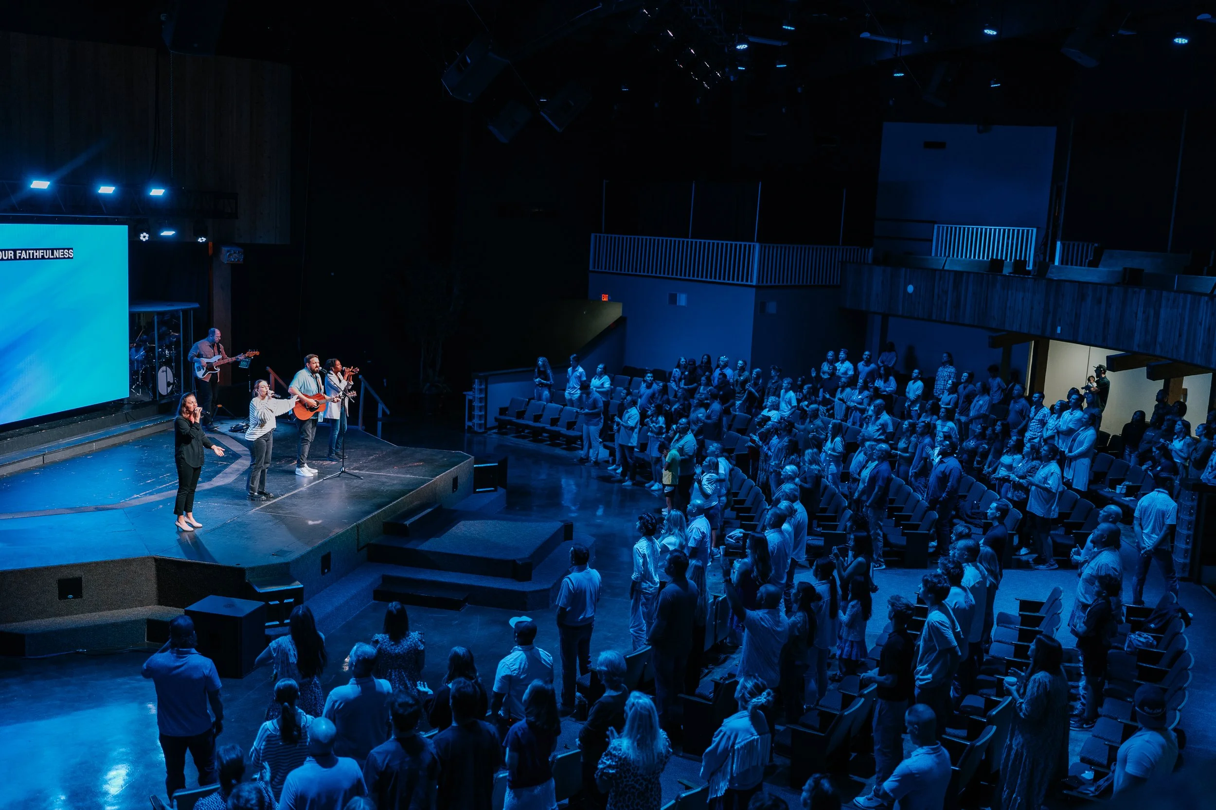 People standing on a stage and in front of it in a dark auditorium, some with hands raised, during a worship service or concert with band playing music.
