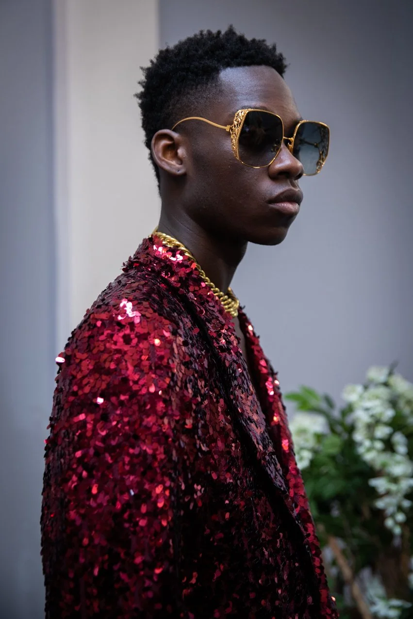 A young person with dark skin and short curly hair wearing oversized gold sunglasses, a shiny red sequined jacket, and a gold chain necklace, standing indoors with a potted plant in the background.