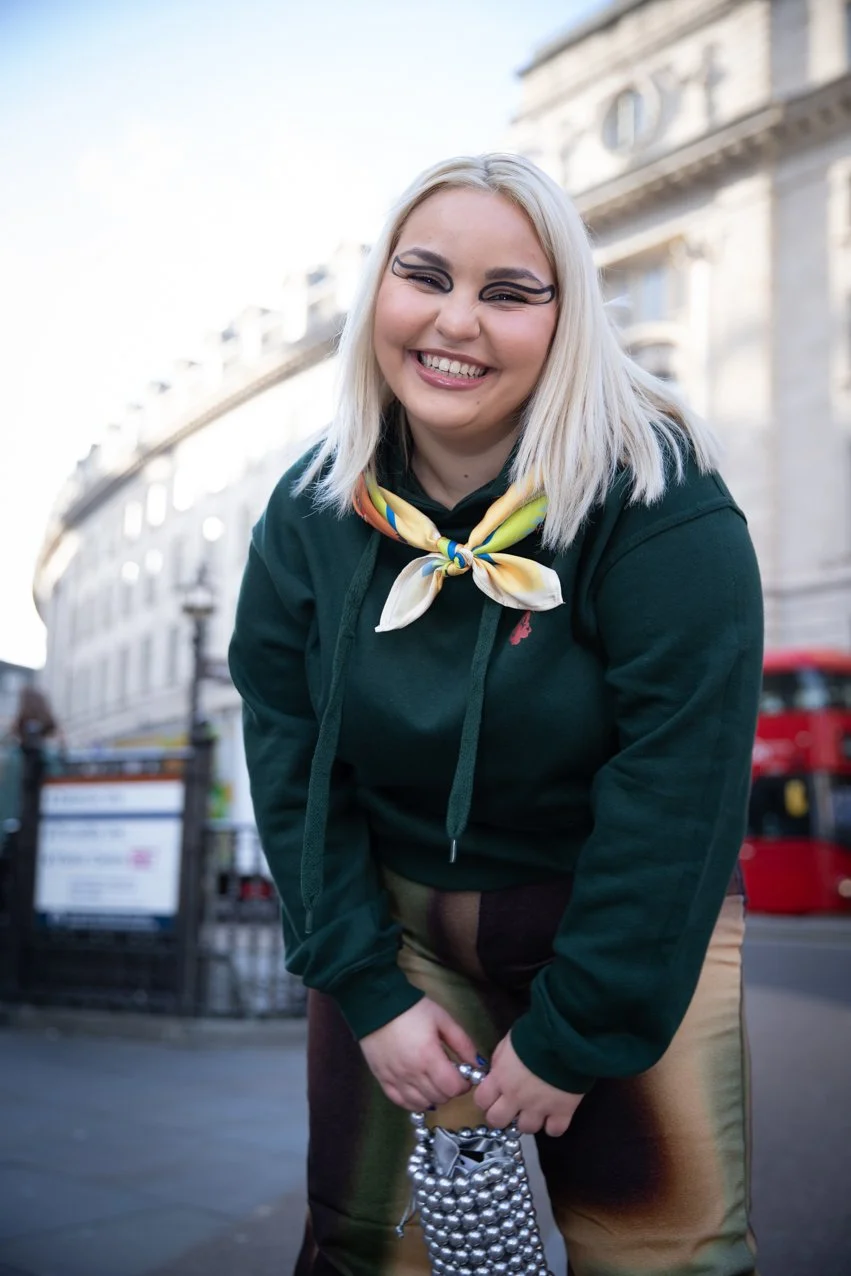 A smiling young woman with platinum blonde hair and makeup, wearing a dark green hoodie, a colorful neck scarf, and patterned pants, standing outdoors on a city street with buildings and a red bus in the background.