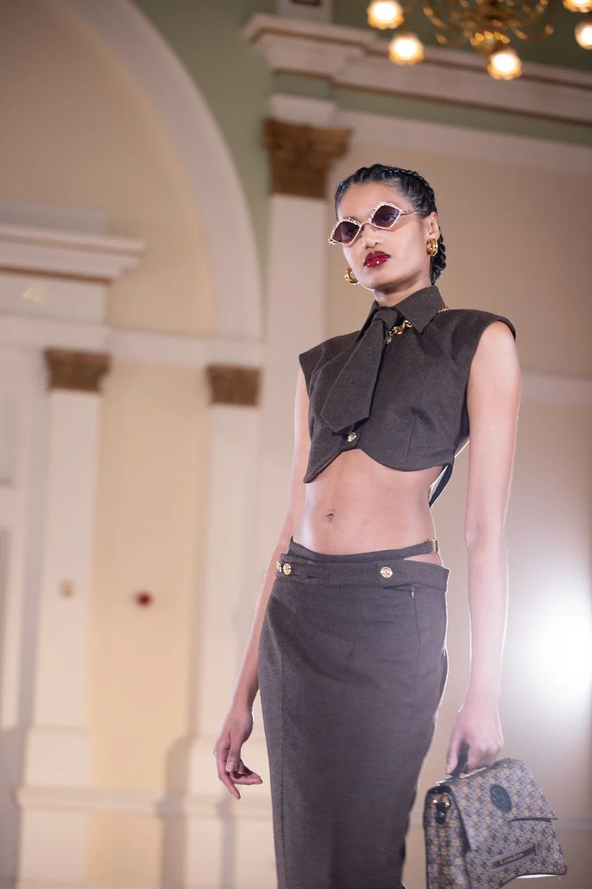 A woman with dark hair styled in braids, wearing sunglasses, red lipstick, gold earrings, a sleeveless dark gray crop top with a bow and gold accents, matching high-waisted pants with gold buttons, and holding a designer handbag, walking in a grand i