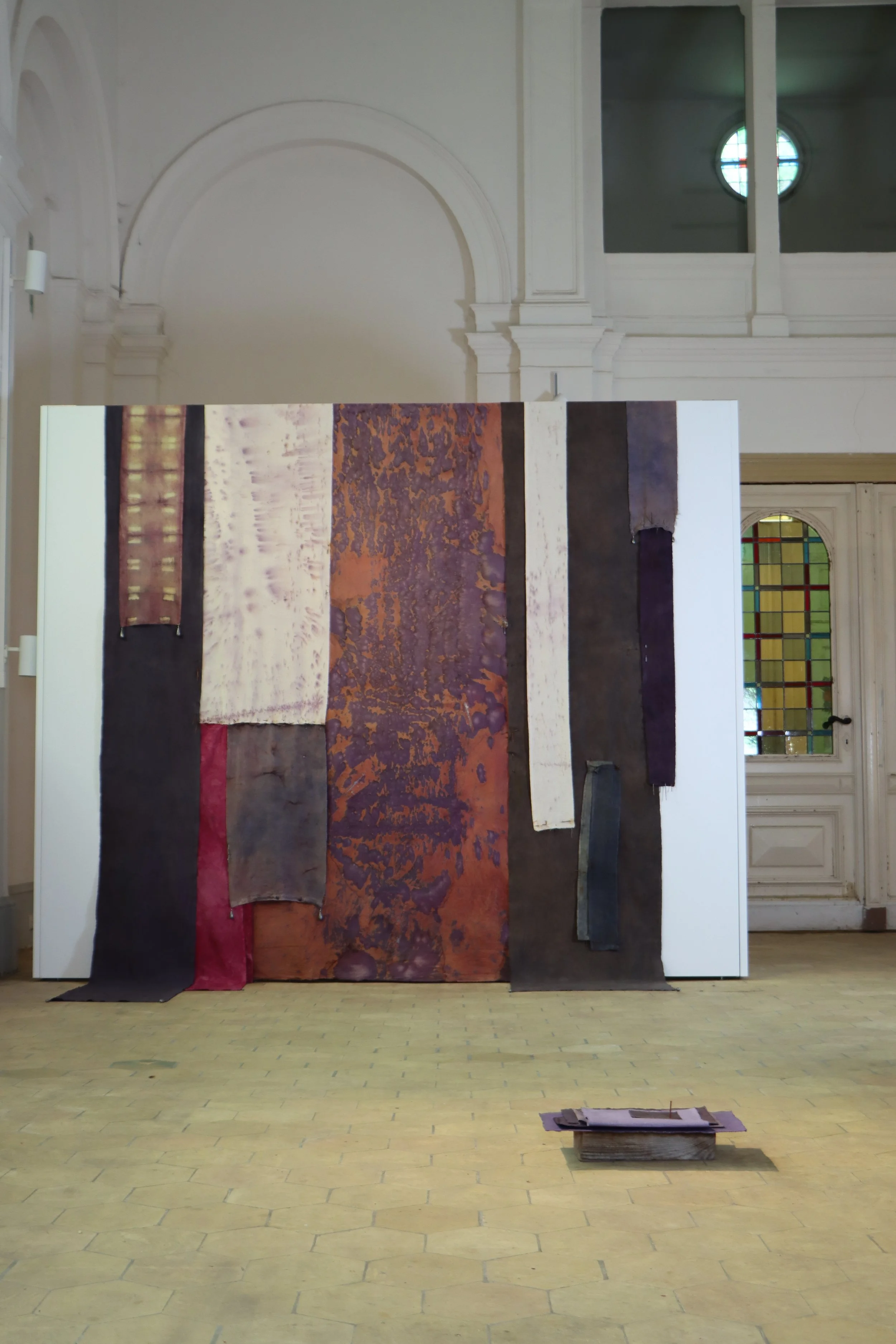 Colorful textile art display with multiple hanging strips of fabric in various colors and textures, arranged vertically on a white wall inside a room with high ceilings and stained glass windows.