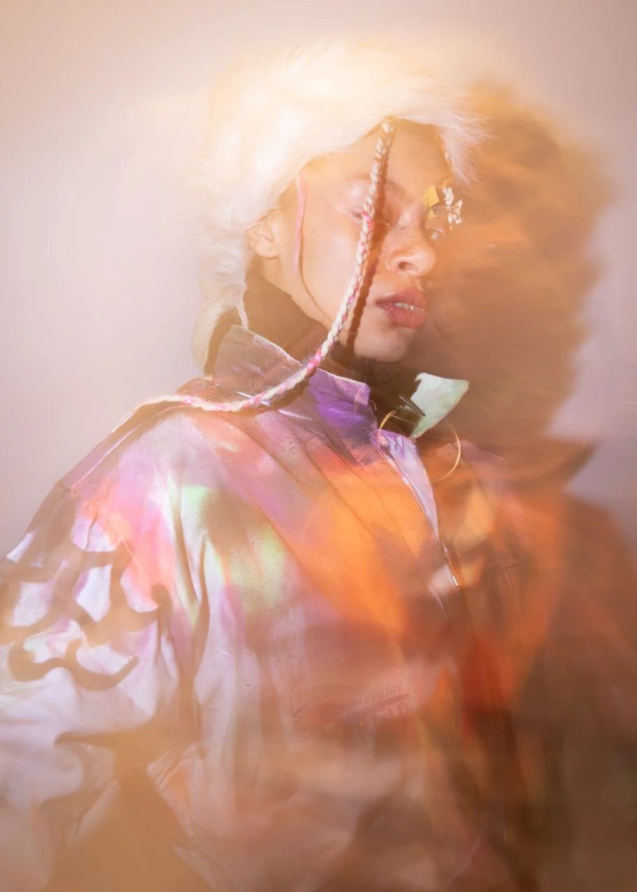 A woman with platinum blonde hair styled in a fluffy manner, wearing colorful clothing, with a dreamy and abstract background.