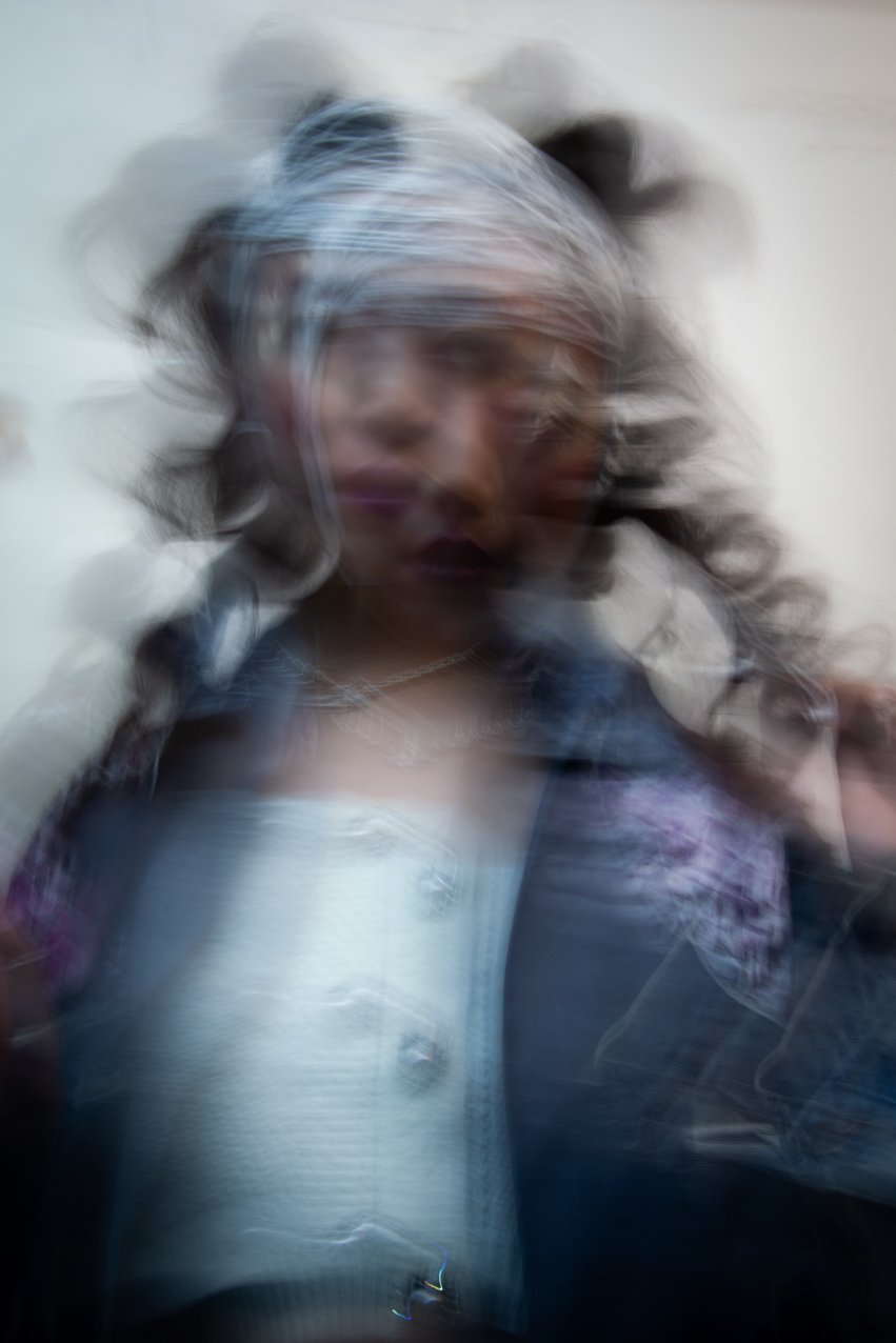 Blurry image of a woman with curly hair, wearing a headscarf, dark lipstick, and a denim jacket.