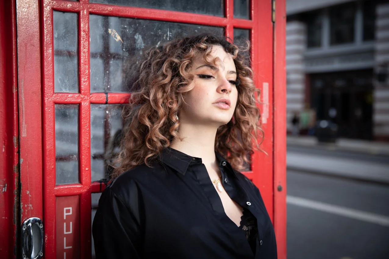 Young woman with curly hair, black shirt, and nose piercing leaning against a traditional red British telephone booth on a city street.