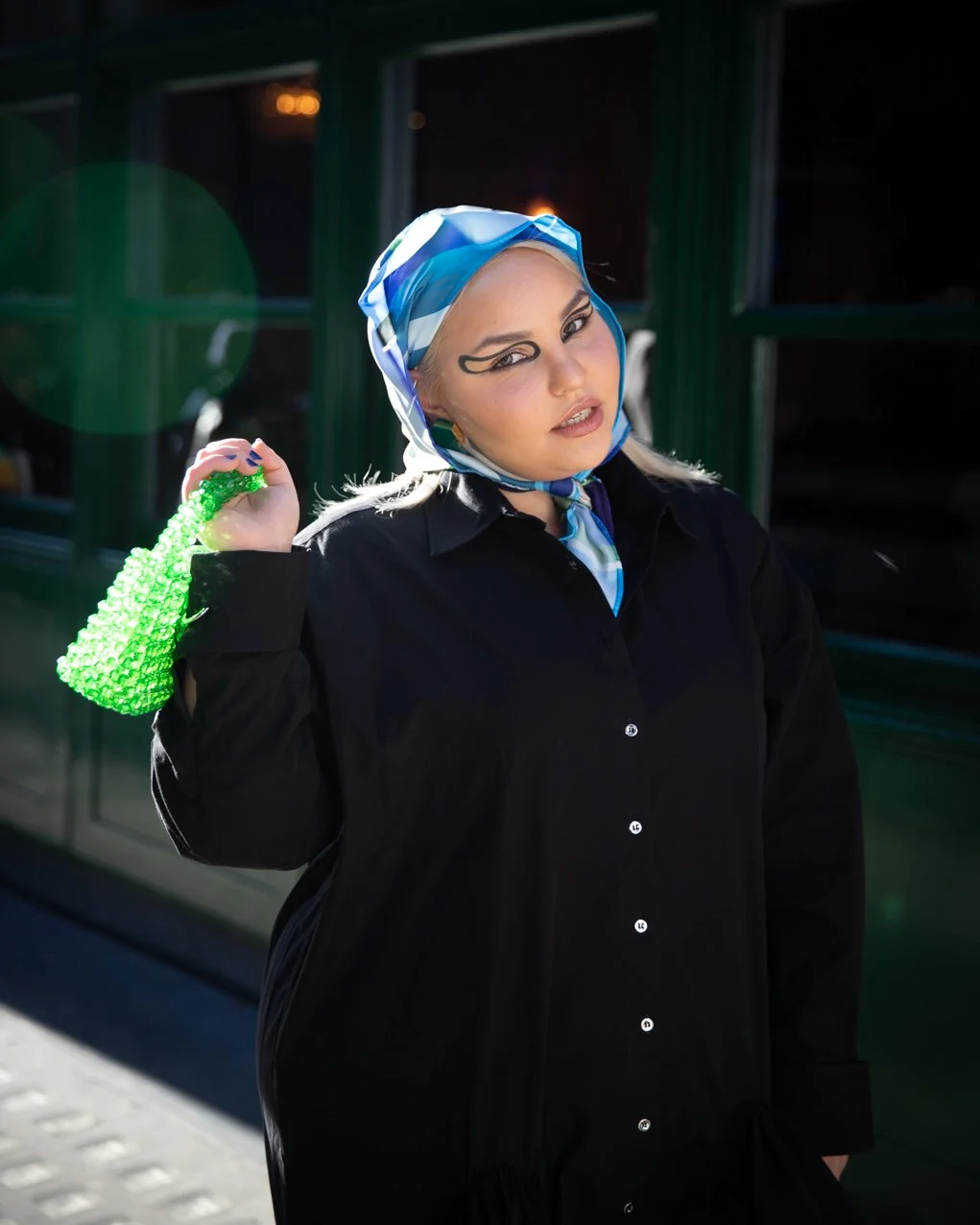 A woman with platinum blonde hair, wearing a blue and white patterned headscarf and bold eye makeup, holds a green beaded purse.