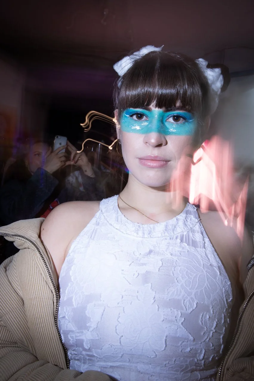 A young woman with brown hair styled in two buns, white bows, and vibrant blue face paint across her eyes, wearing a sleeveless white lace top and a light-colored jacket, standing in a dimly lit room with a blurry background. In the background, someo