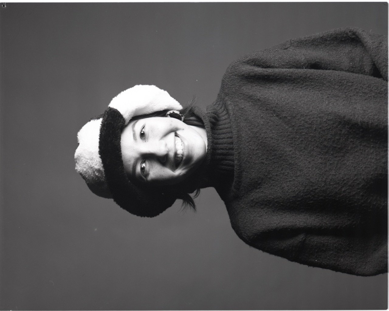 A person smiling, wearing a cozy sweater and a fuzzy hat, against a plain background.