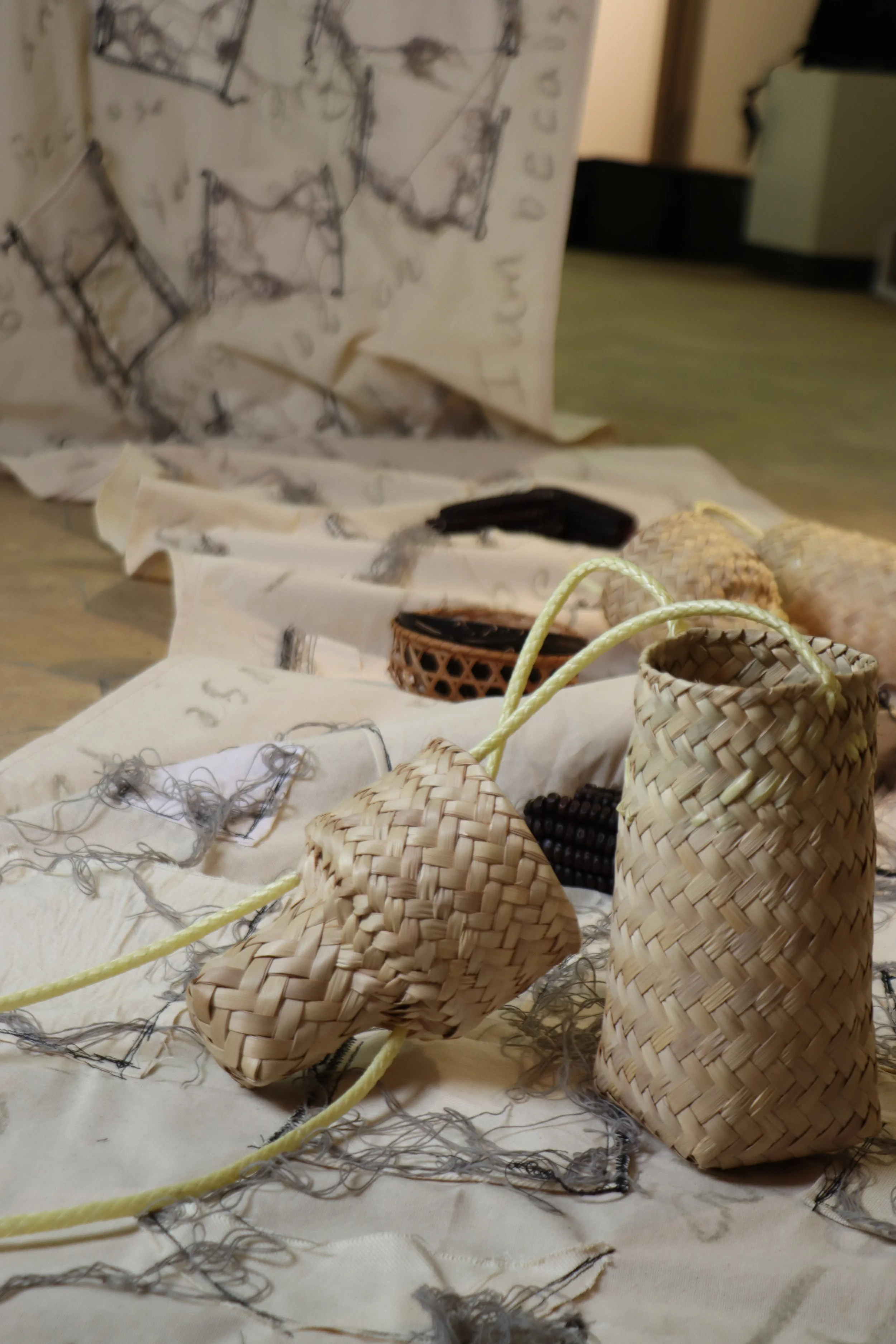 Close-up of woven straw slippers and small baskets on fabric with drawn sketches and threads, with a box in the background.