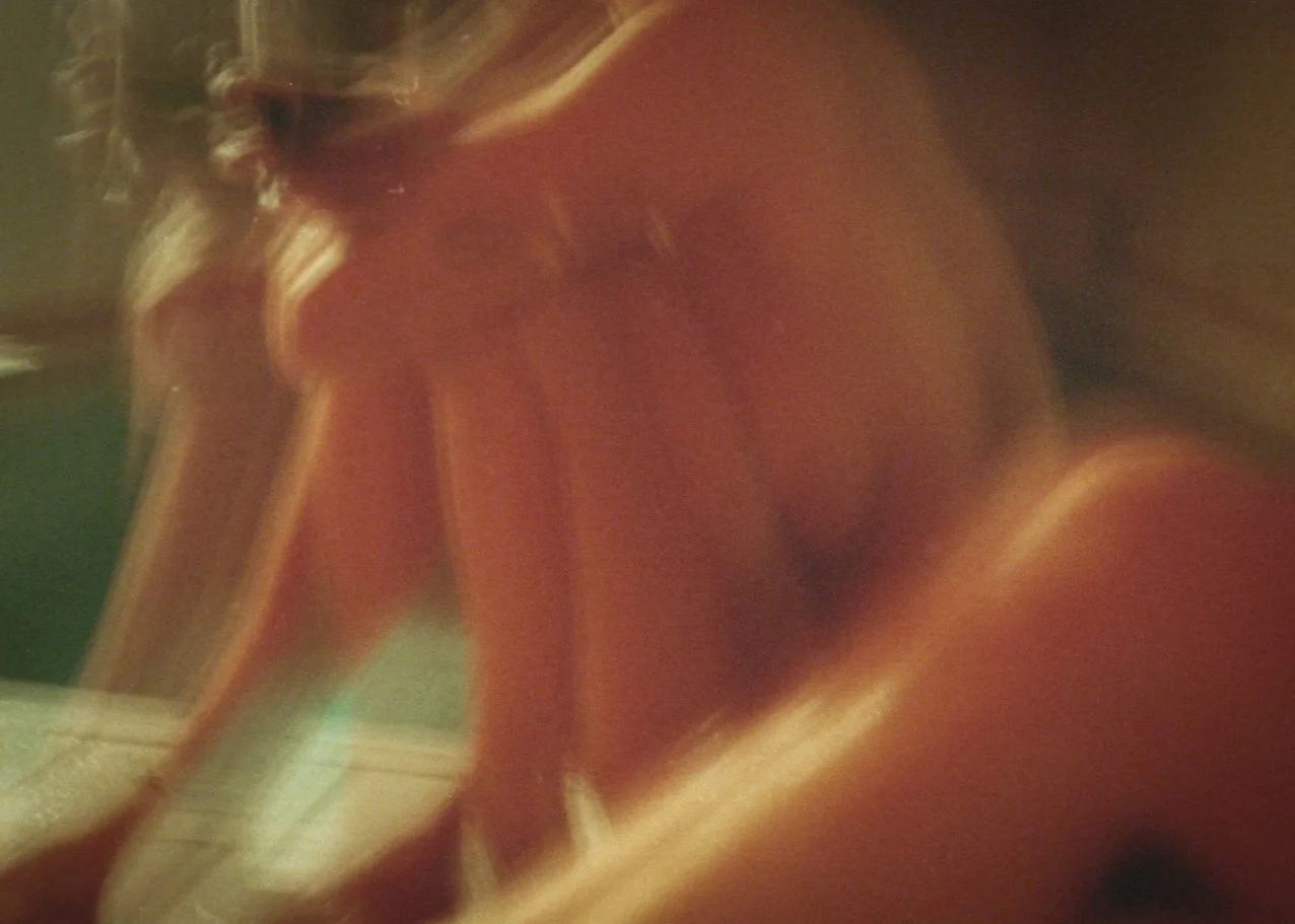 Blurry close-up of a woman with long, wavy blonde hair, wearing a red top or dress, sitting with her knees drawn up.