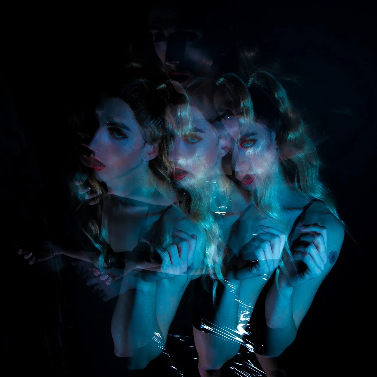 Multi-exposure photograph of a woman with wavy blonde hair, dark eye makeup, and red lipstick, posing against a dark background with blue lighting, creating overlapping images of her face and upper body.