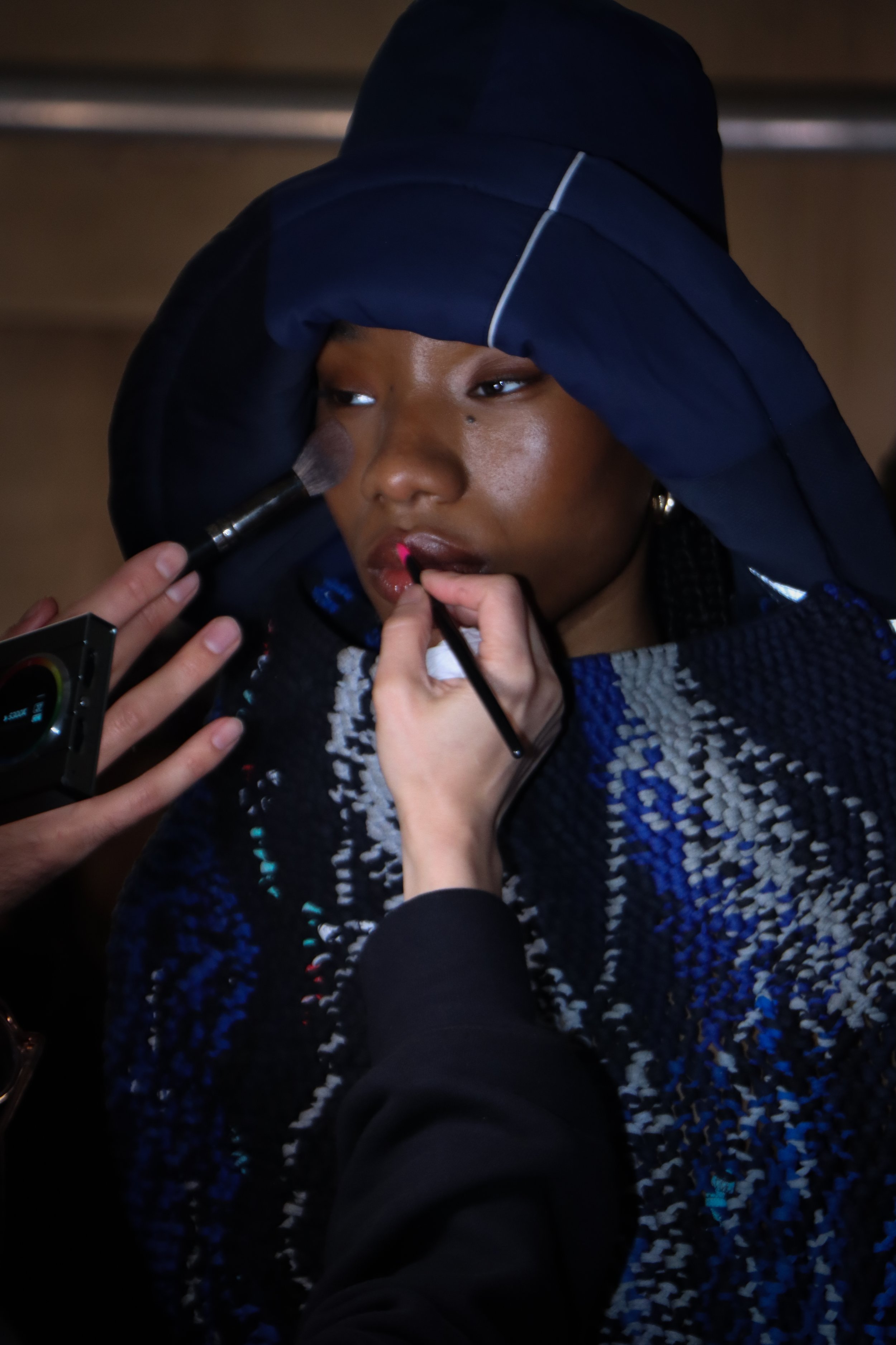 A woman is having her makeup applied. She is wearing a dark head covering and a blue patterned garment. A makeup artist is using a brush to apply lipstick and another person is using a makeup brush on her face.