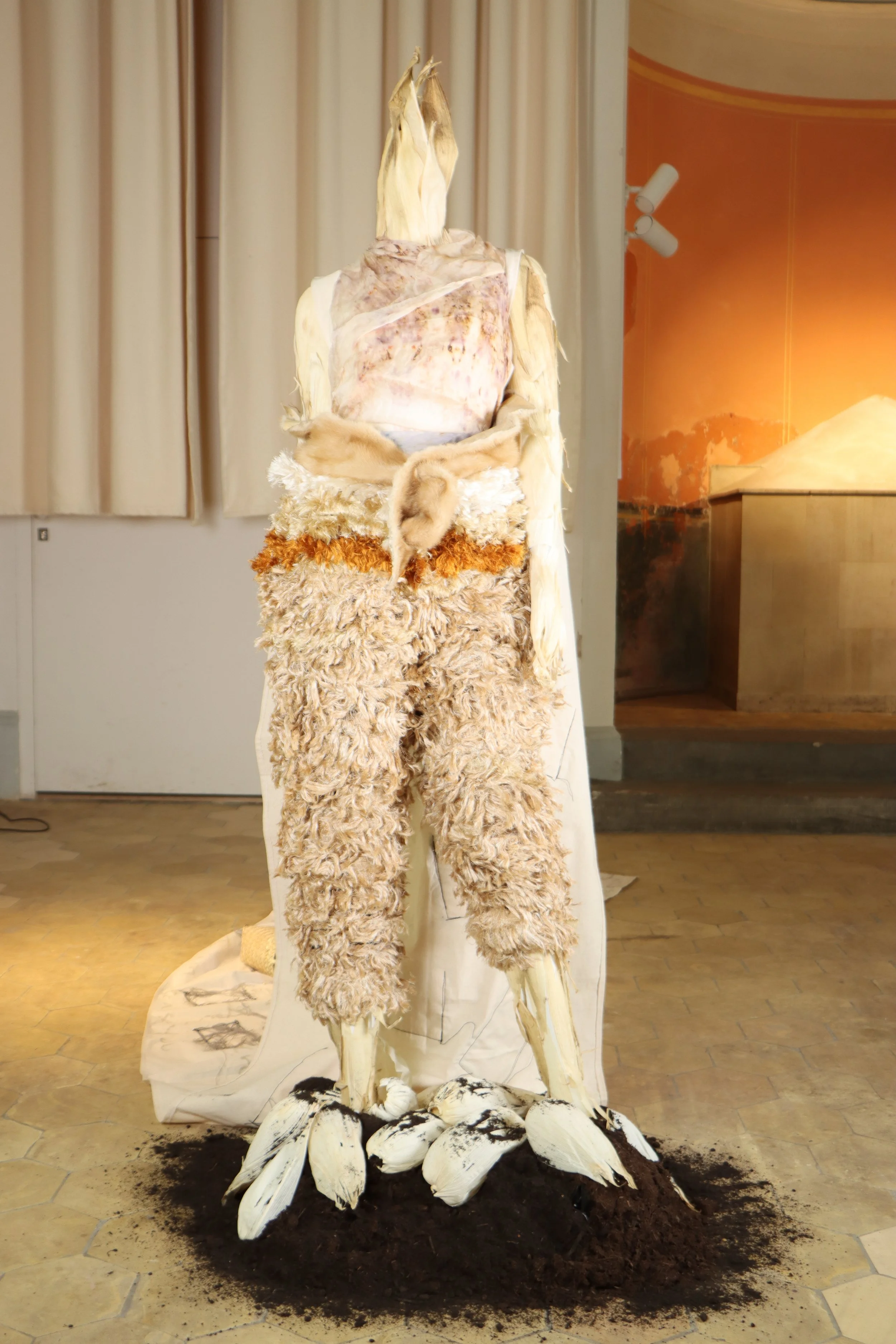 A mixed media art piece resembling a humanoid figure with textured, furry clothing, standing on a soil base surrounded by white, shell-like objects.