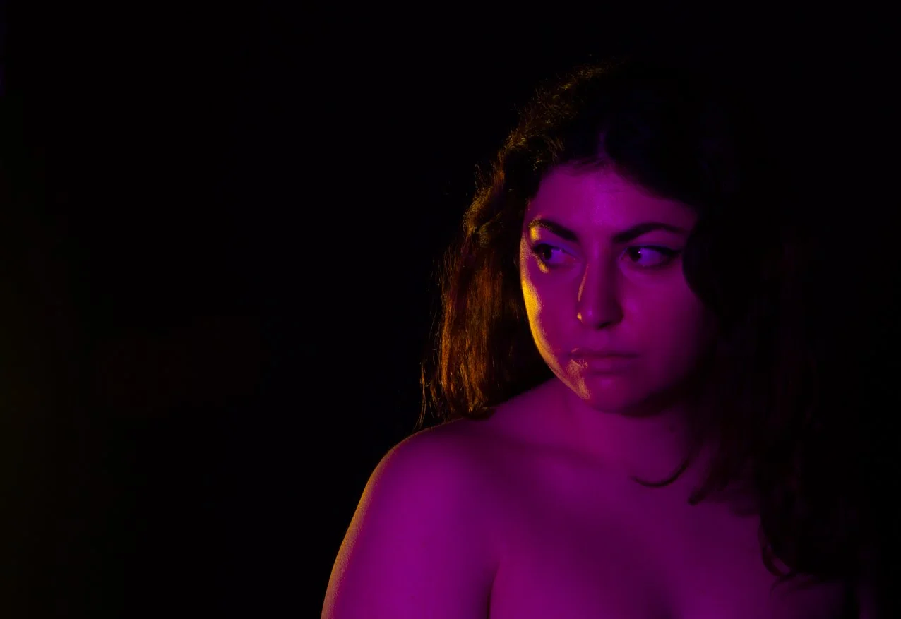 A woman with dark, curly hair is illuminated with purple and yellow lighting against a black background, looking to her right.