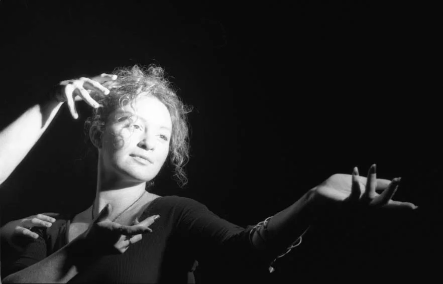 A woman with curly hair and a serious expression extends her arms and hands in expressive gestures, with dramatic lighting creating high contrast in a black background.