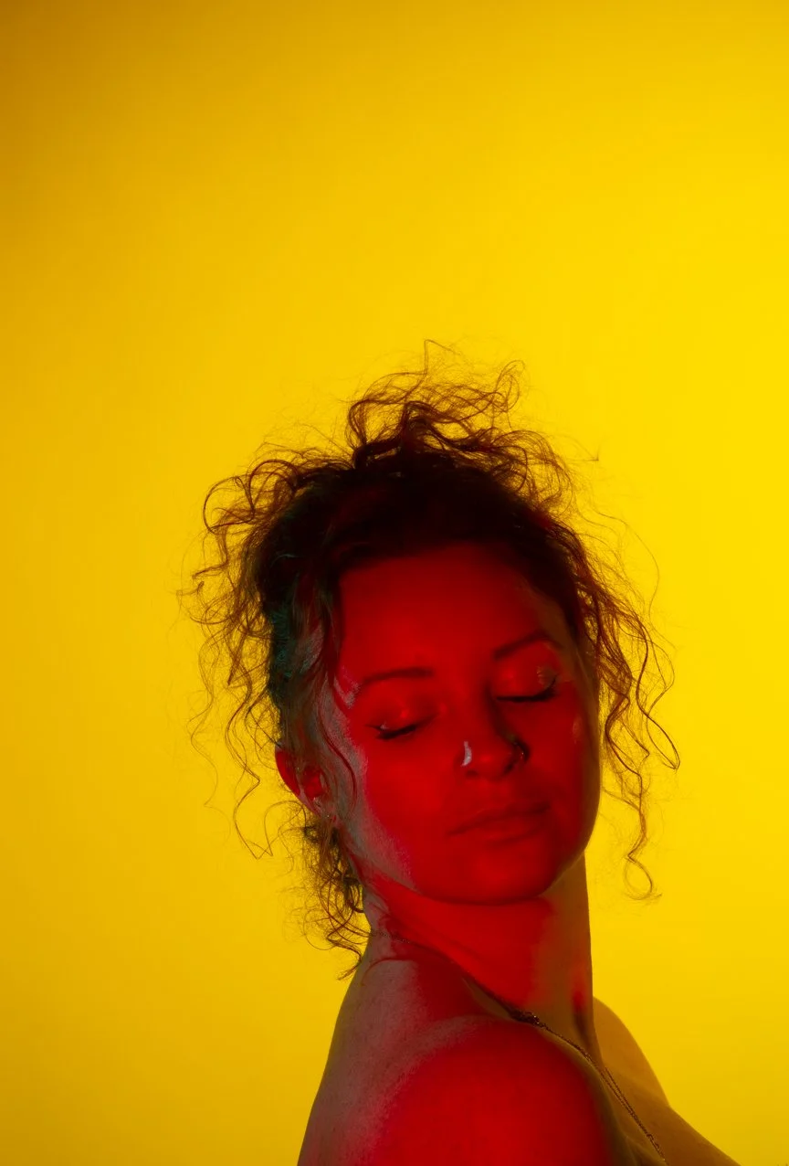 Portrait of a woman with curly hair and closed eyes against a yellow background, illuminated by red and green lighting.