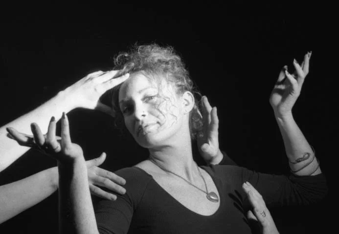 Black and white photo of a woman with curly hair, smiling softly, with multiple hands reaching towards her face and shoulders.