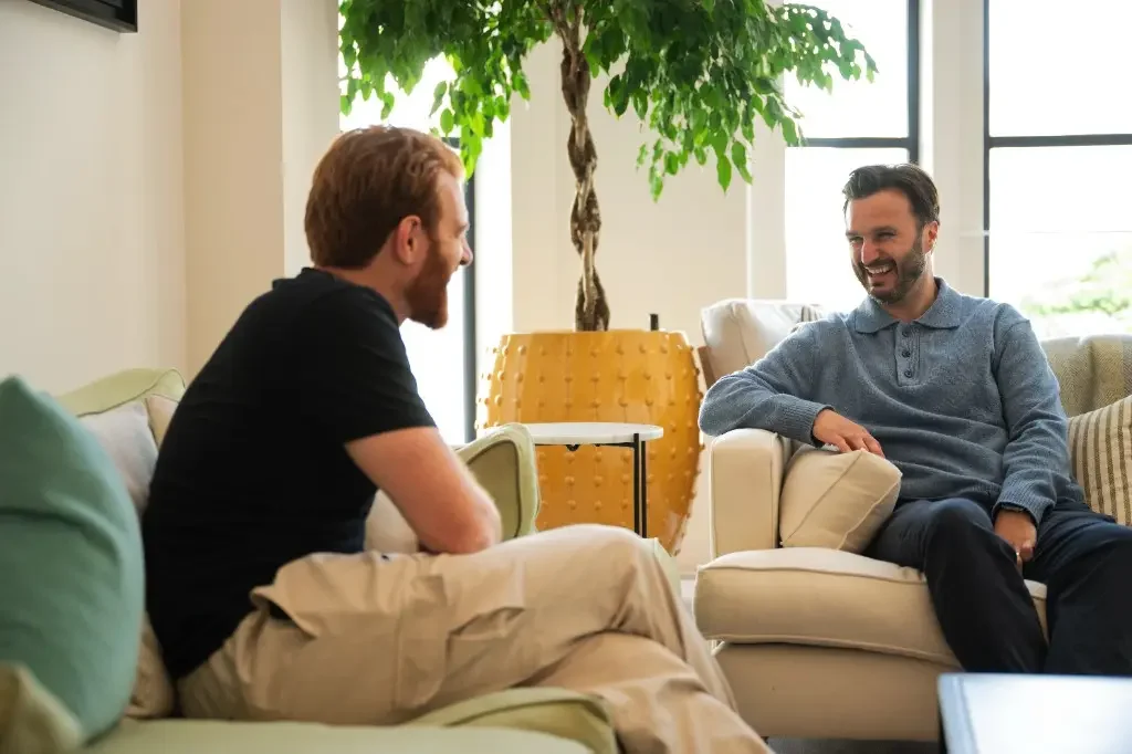 Two men sitting on a sofa in a cozy, well-lit room, engaging in a friendly conversation and smiling.