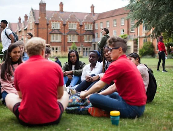 Group of diverse teenagers sitting on grass and talking on a college campus lawn with historic red brick buildings in the background.