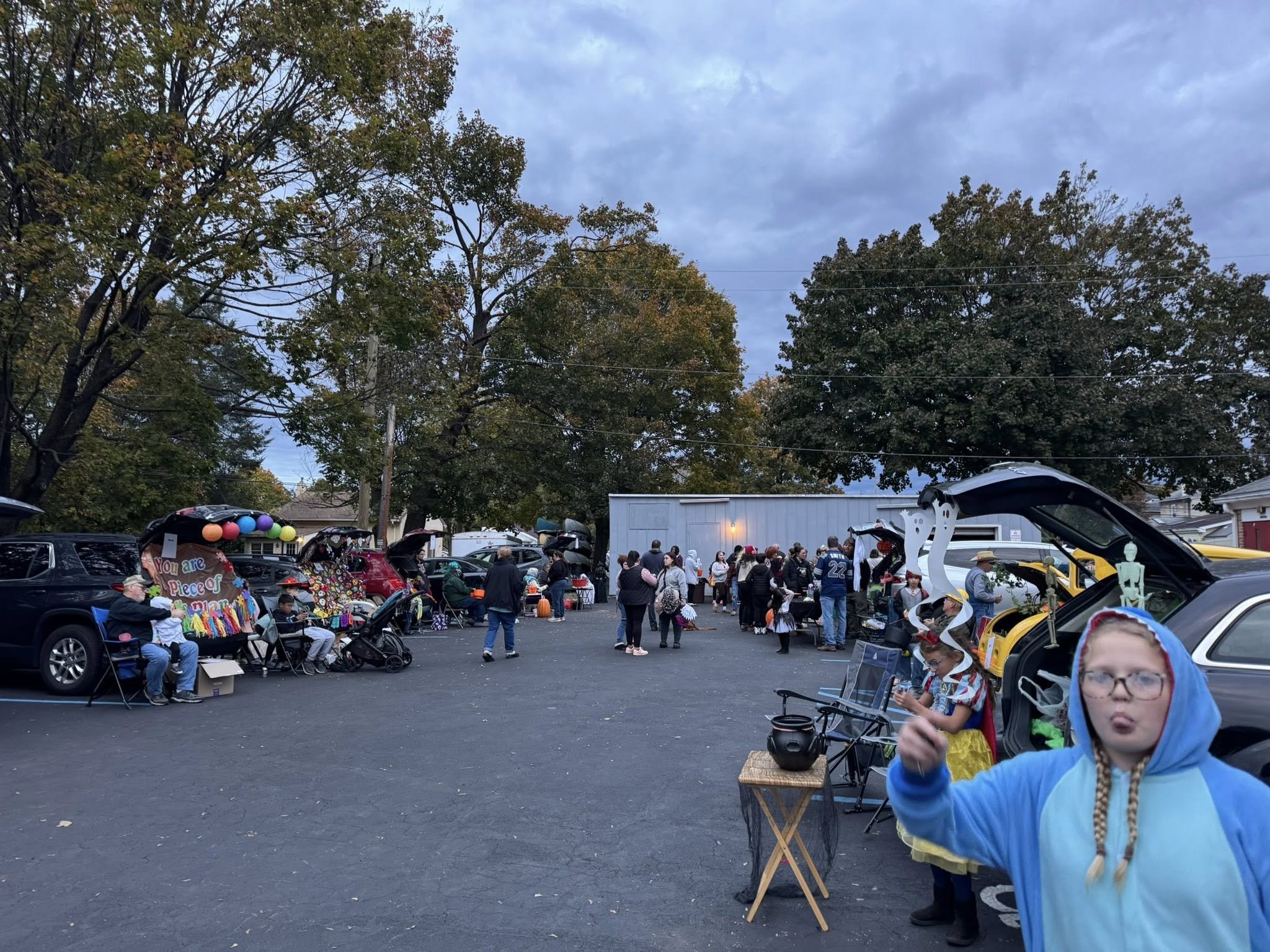 A community gathering in a parking lot with trees and cloudy sky in the background. People are socializing, and there are cars, tents, and decorations, including colorful lanterns. Children and adults are present, some seated and some standing.